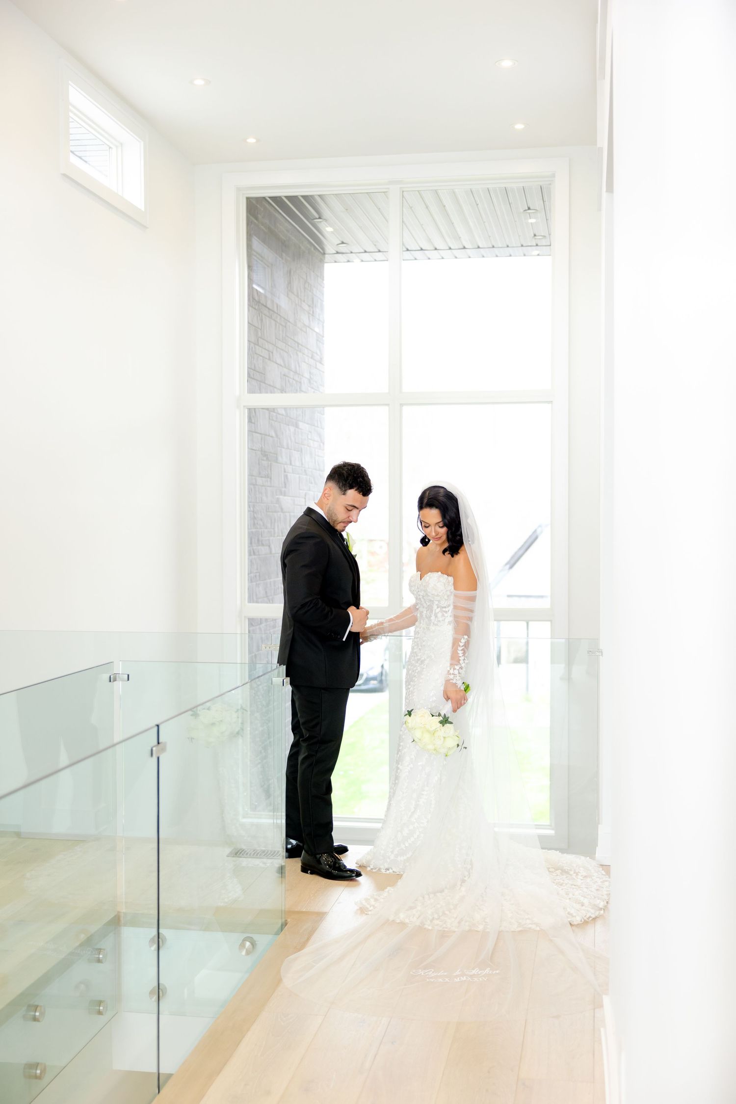 A wedding couple stands in an elegant white hallway with glass railings and natural light streaming in.
