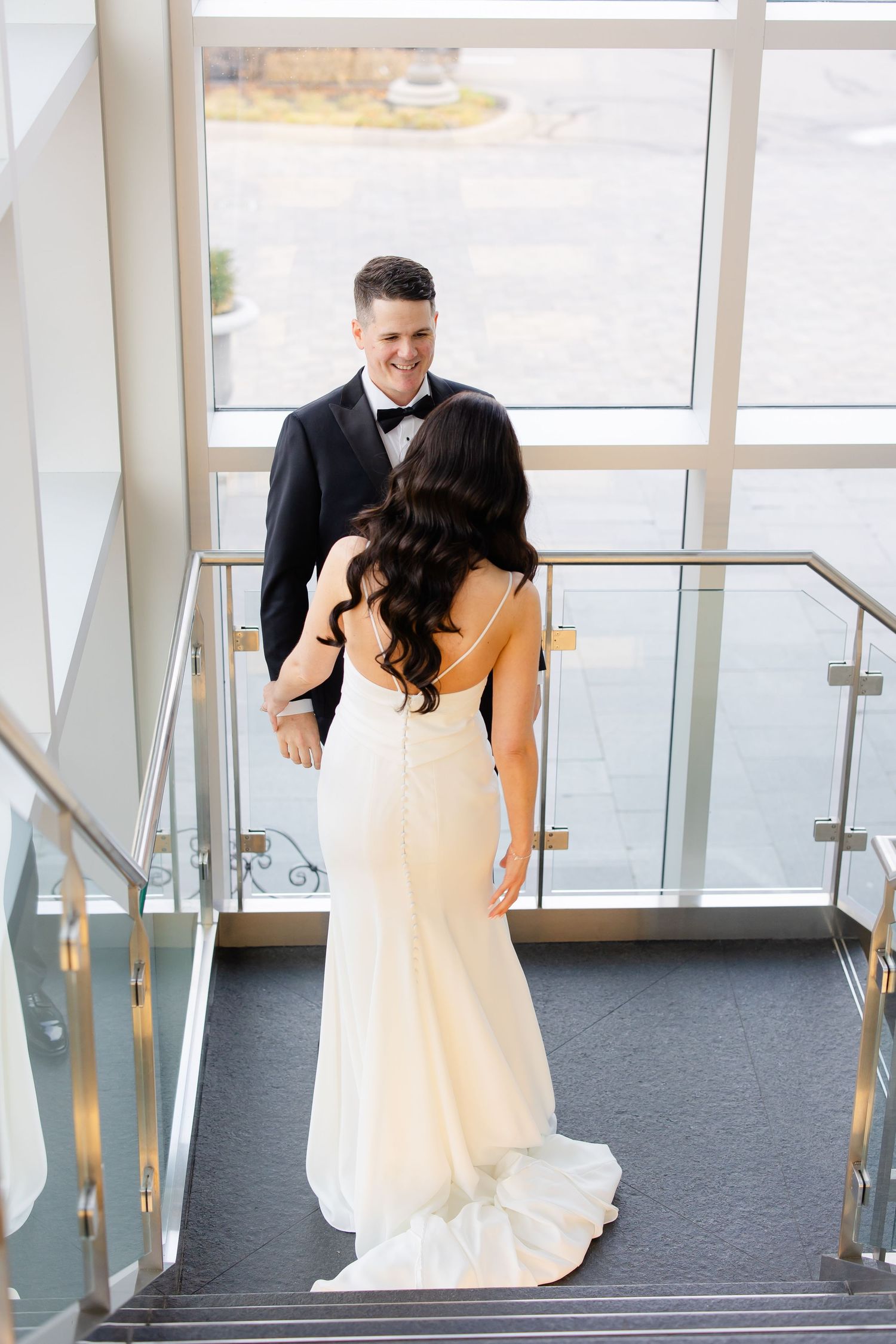 Bride in flowing white dress stands on modern staircase while groom looks on from above.