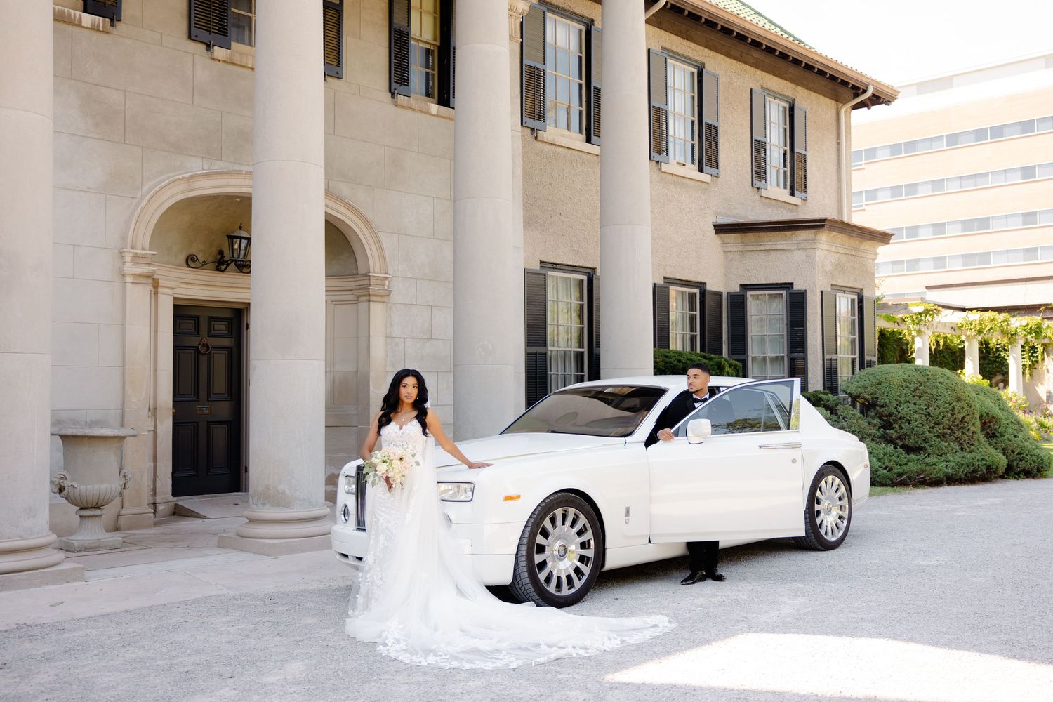 A bride in a white lace gown poses next to a pristine white Rolls-Royce at an elegant venue.