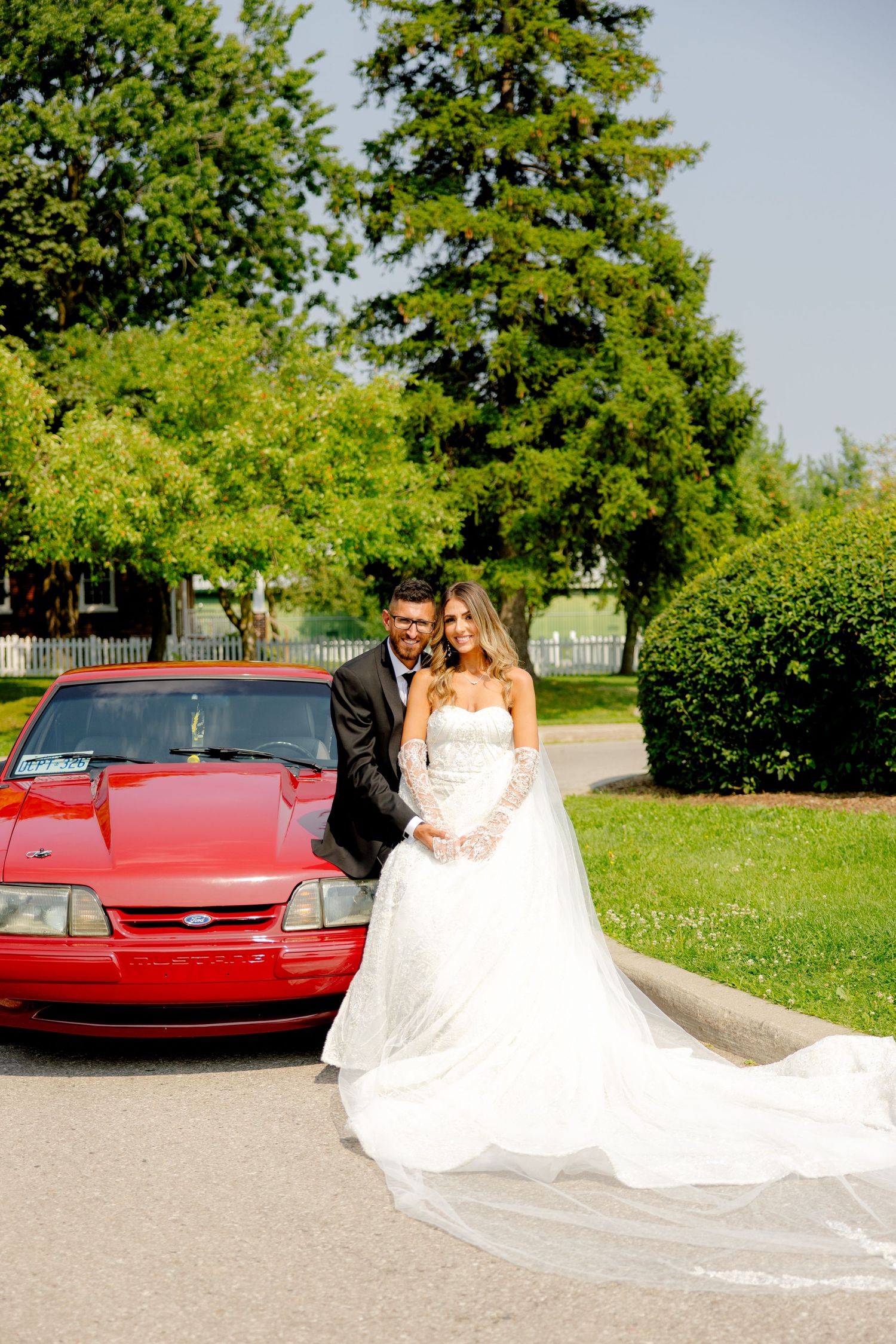 Intimate wedding portrait with couple posed by their red car in a serene park setting.