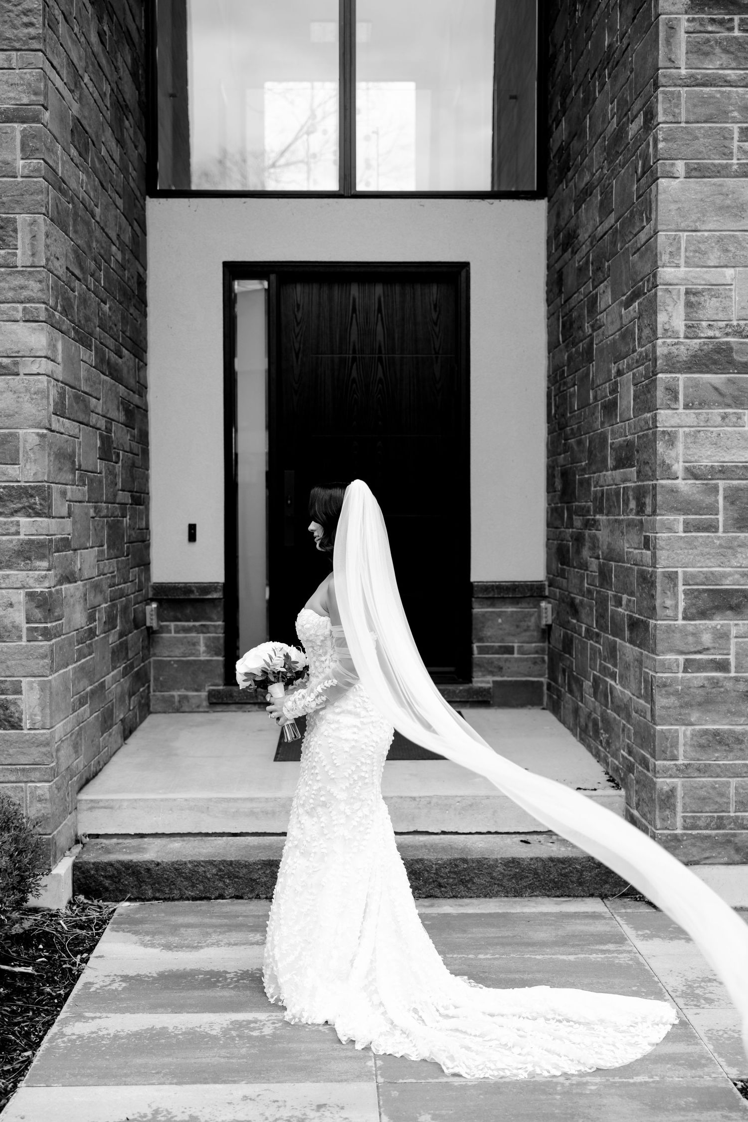 A bride in a lace gown with flowing veil stands in a doorway holding a white bouquet.