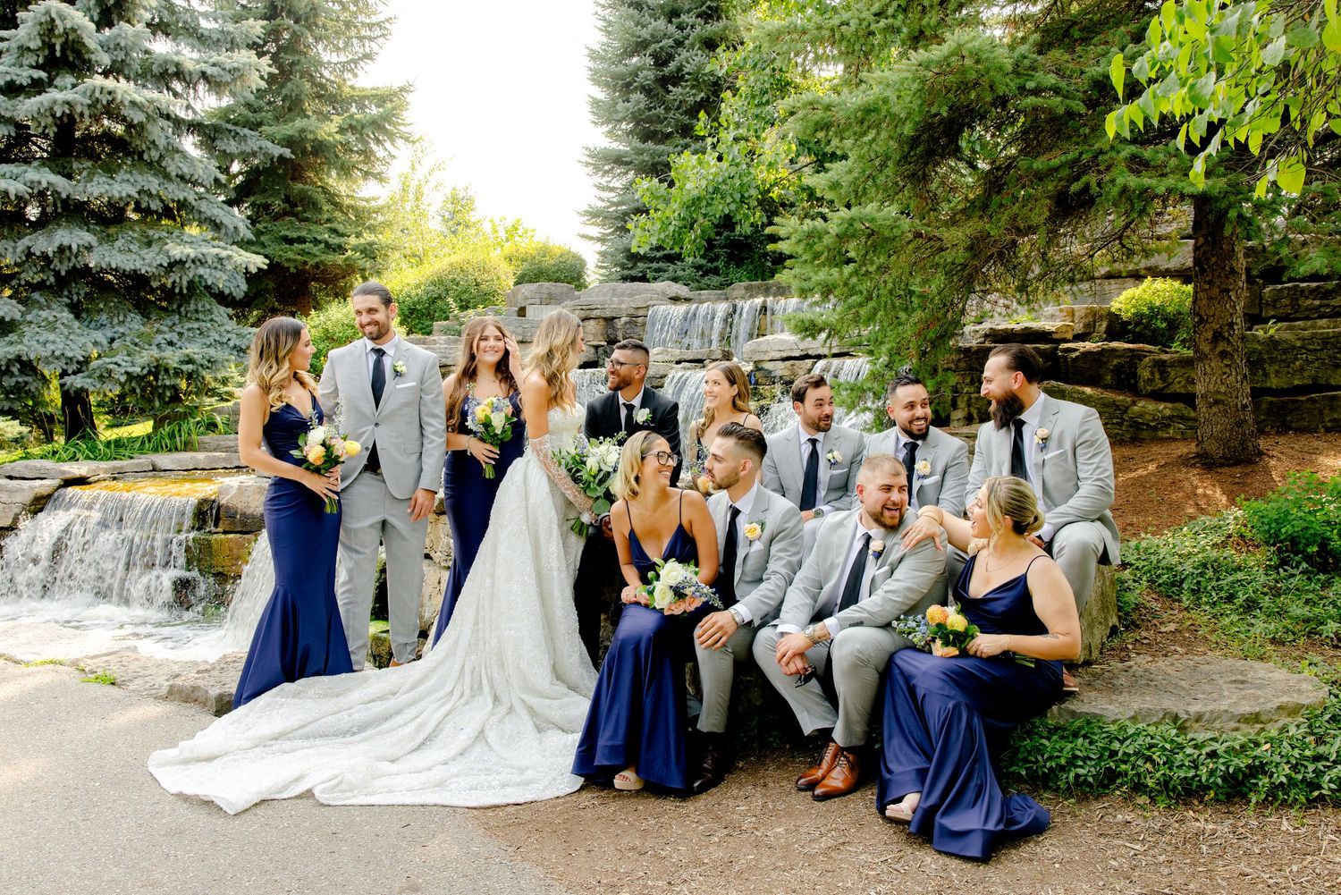 Joyful wedding party group shot with bridesmaids in navy and groomsmen in gray by waterfall.