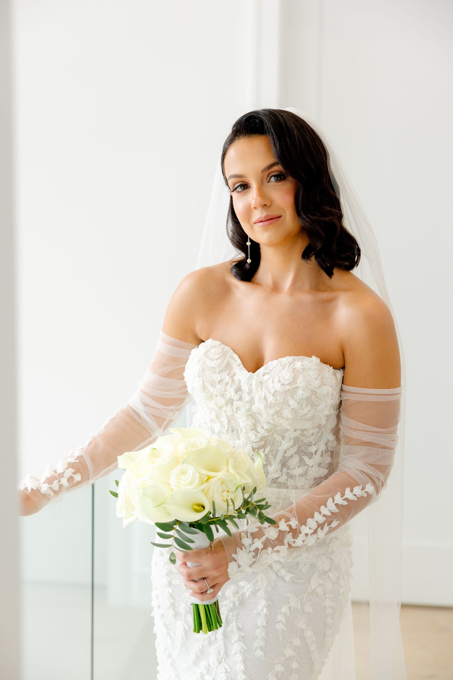An intimate bridal portrait featuring a strapless lace gown with sheer sleeves and white rose bouquet.