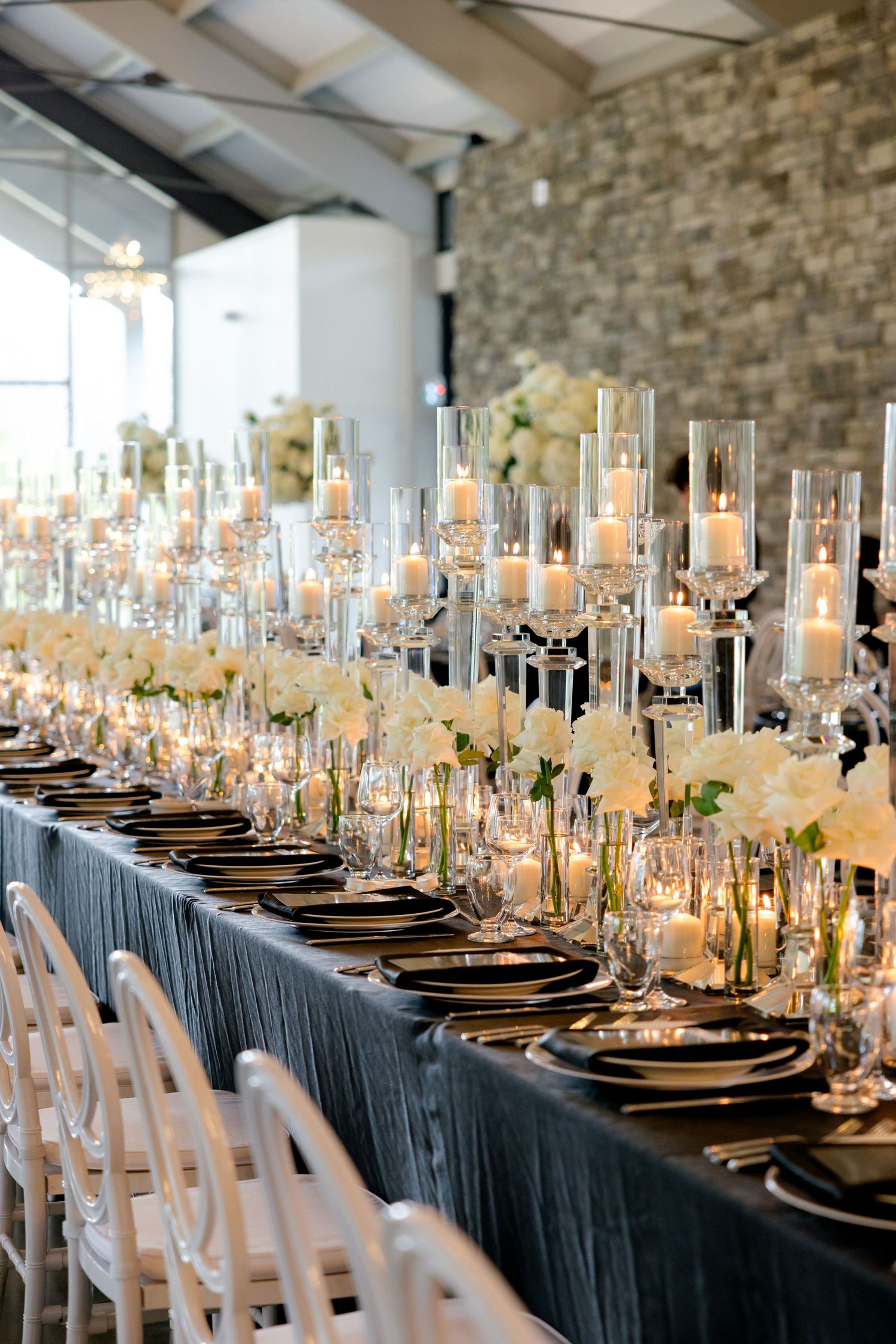 Long wedding reception table with crystal candleholders and white chairs against a stone wall.