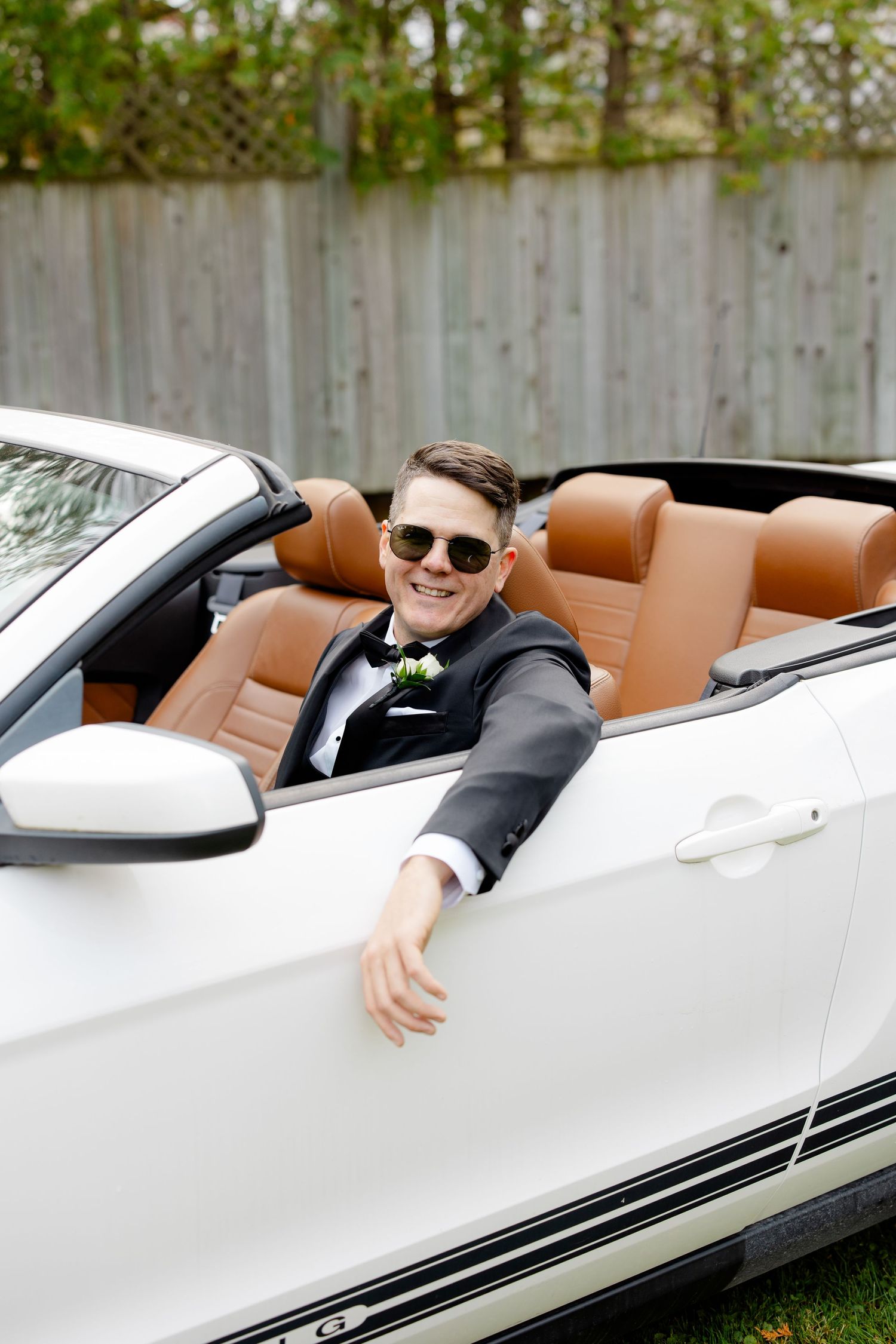 A person in a tuxedo smiles while sitting in a white convertible car with tan leather interior.