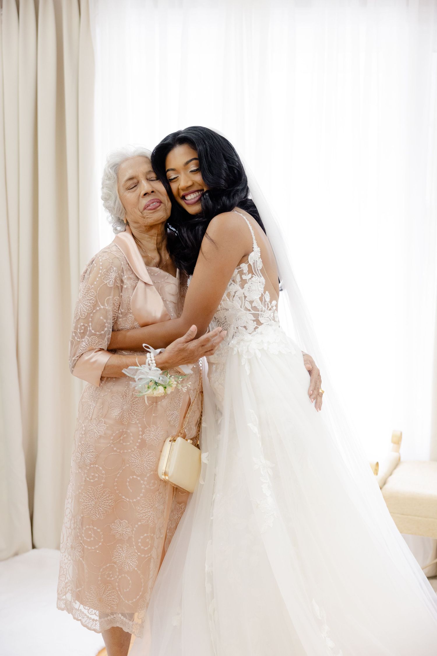 Tender embrace shared between bride in white lace gown and wedding guest in blush dress.