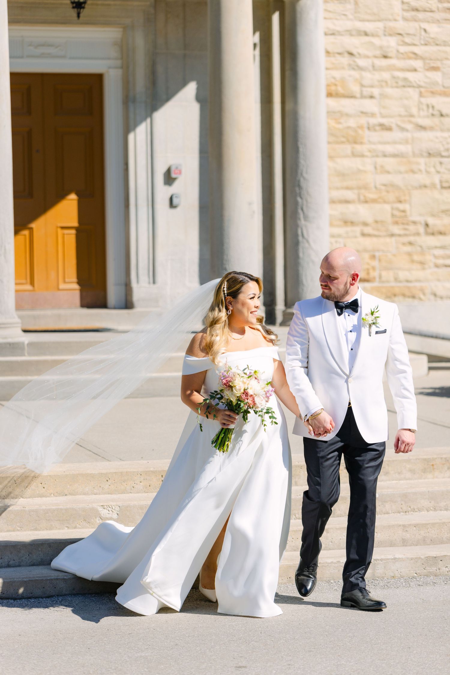 Couple in wedding attire walking together outside elegant stone building with columns.