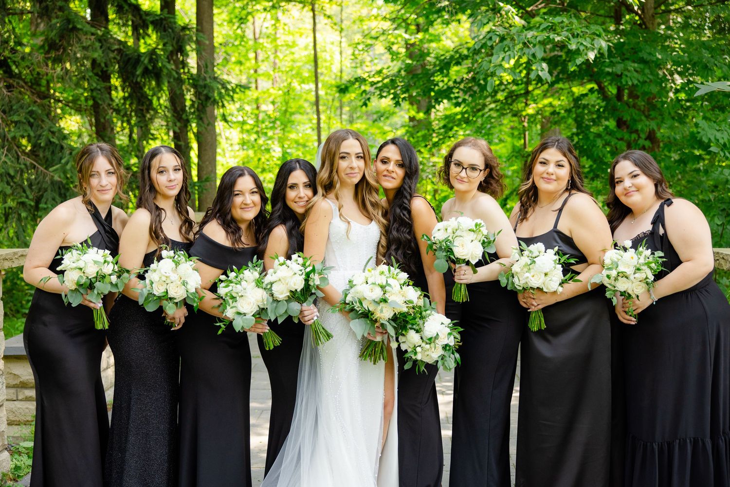 Bridal party in elegant black gowns and white dress gather together holding matching white floral bouquets.