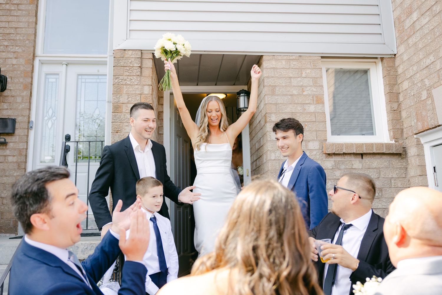 Newlyweds celebrate on steps of brick house as wedding guests applaud below.