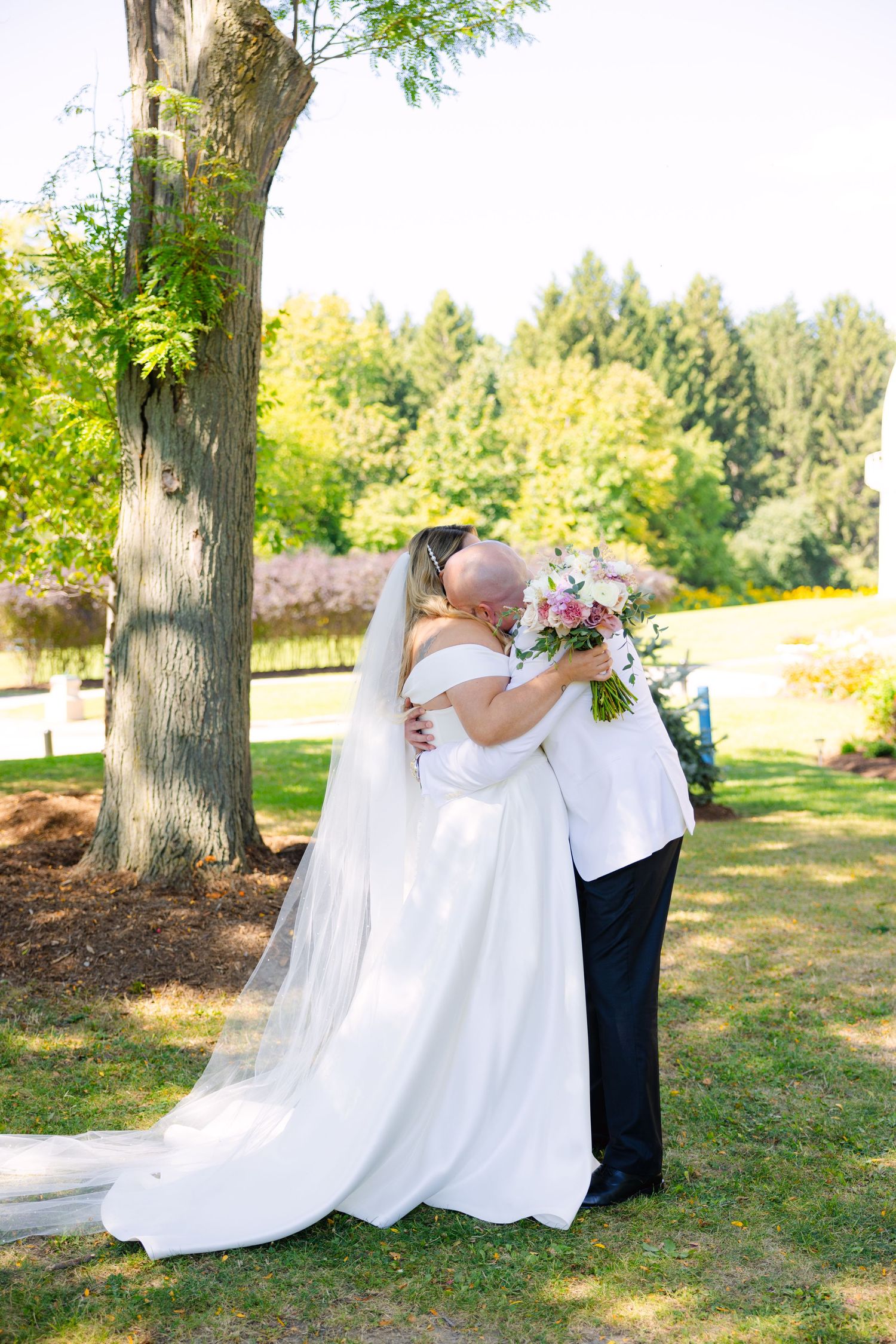 Intimate wedding embrace under a large tree with bride's flowing white dress trailing behind.