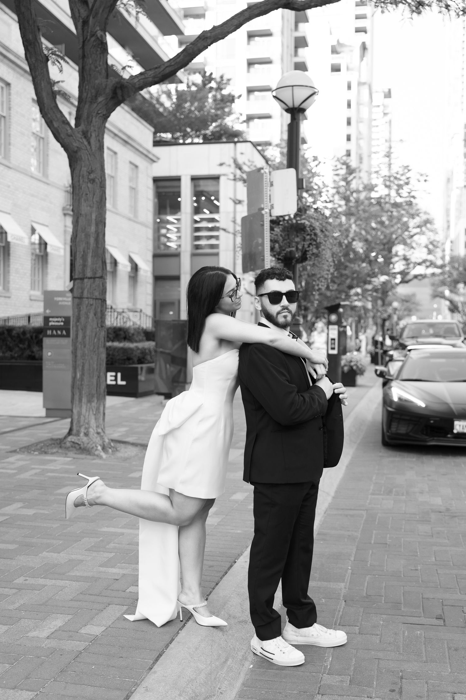 Couple in formal attire poses playfully on city sidewalk with trees and buildings in background.