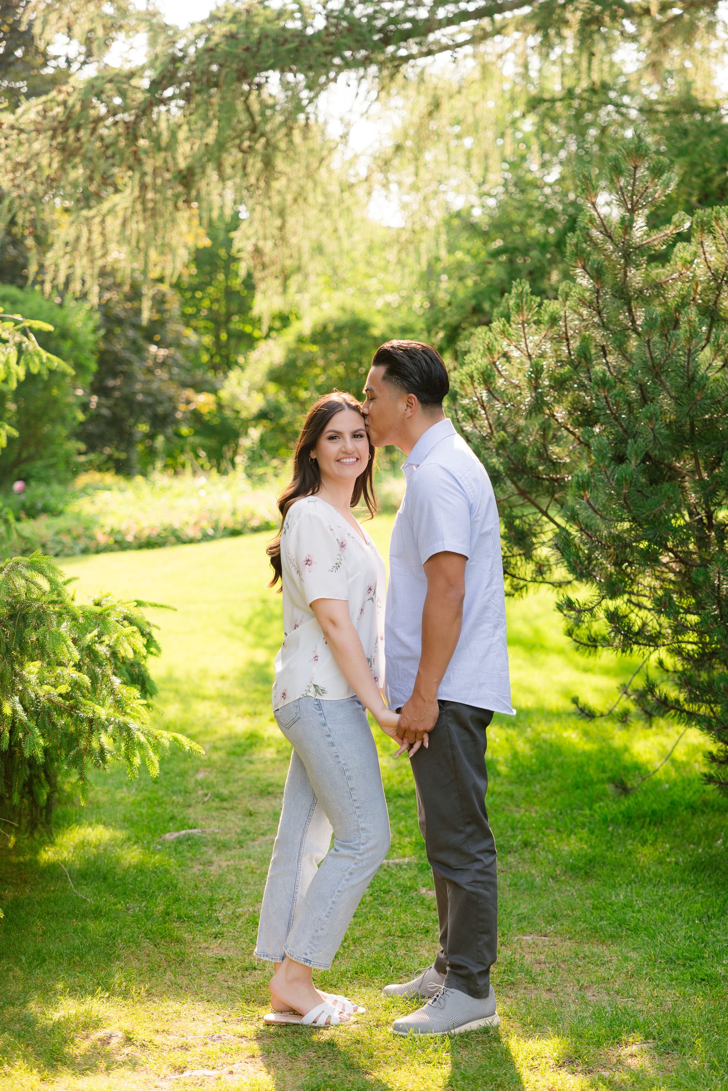 A couple shares an intimate moment while standing among evergreen trees in a sunlit garden setting.