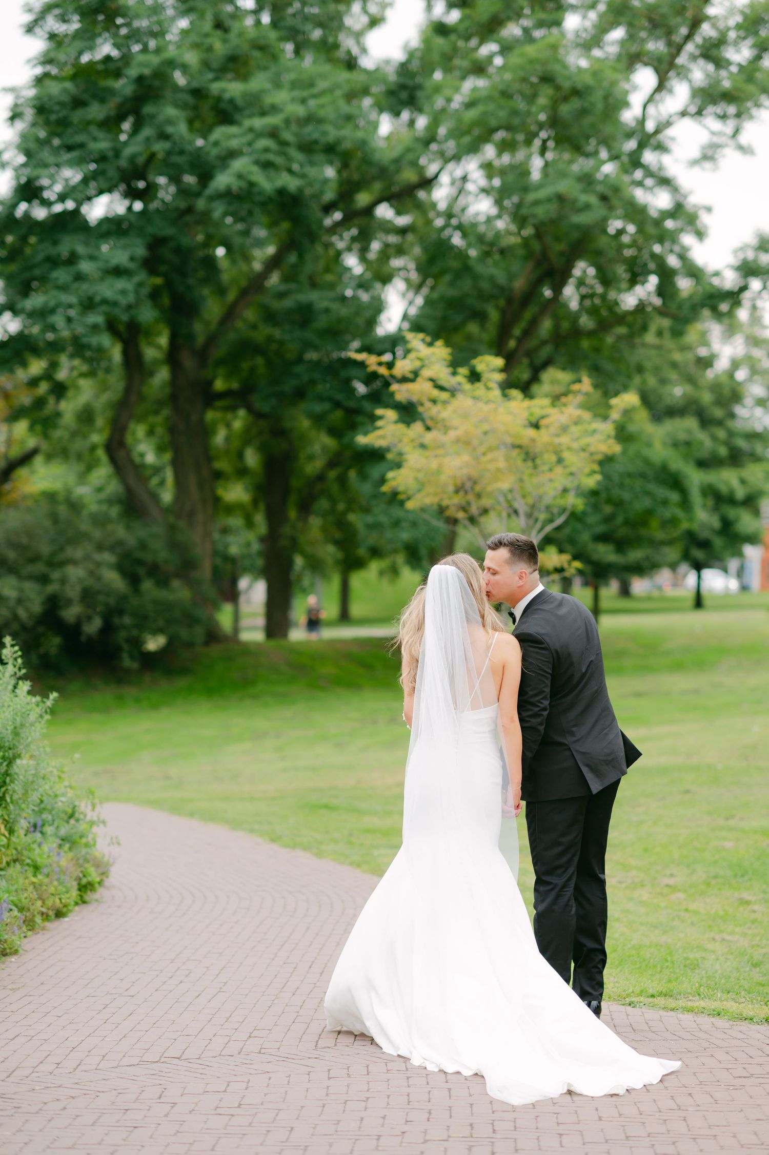 Newlyweds share an intimate moment on a tree-lined path in a lush garden setting.