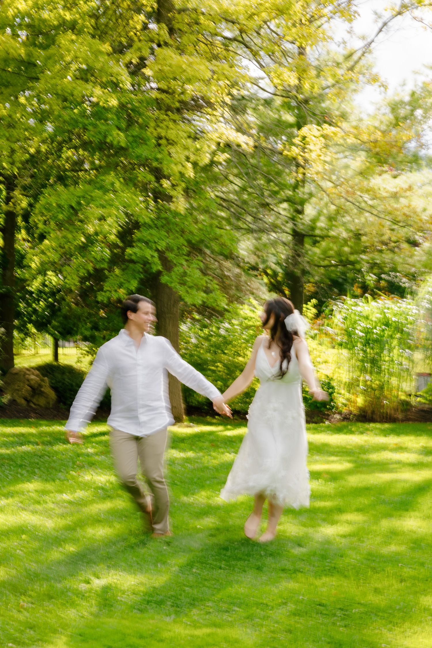 A couple in wedding attire runs playfully through a sunlit garden with bright green foliage.
