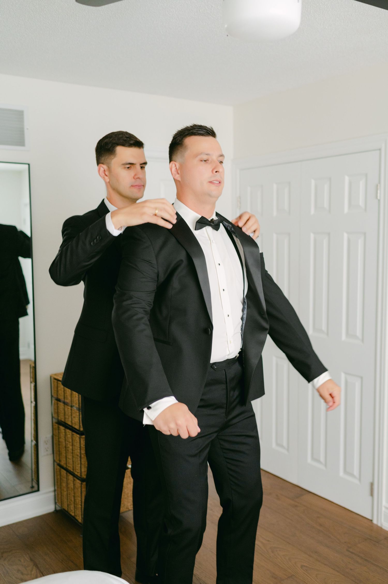 Two people in tuxedos preparing for a formal event in a bright indoor setting.