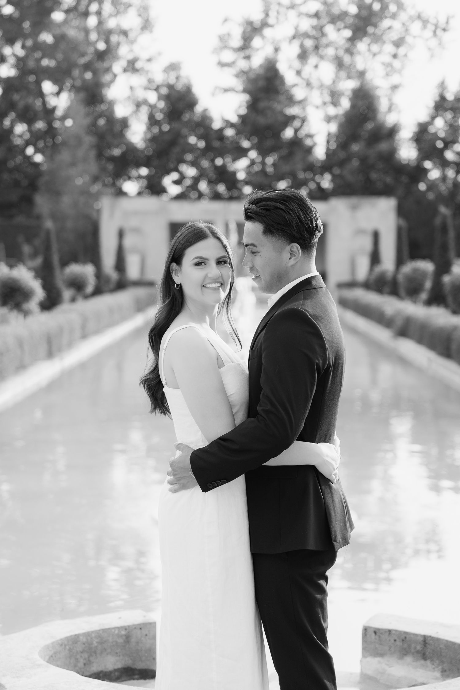A black and white portrait of a couple embracing by a reflecting pool in a formal garden.