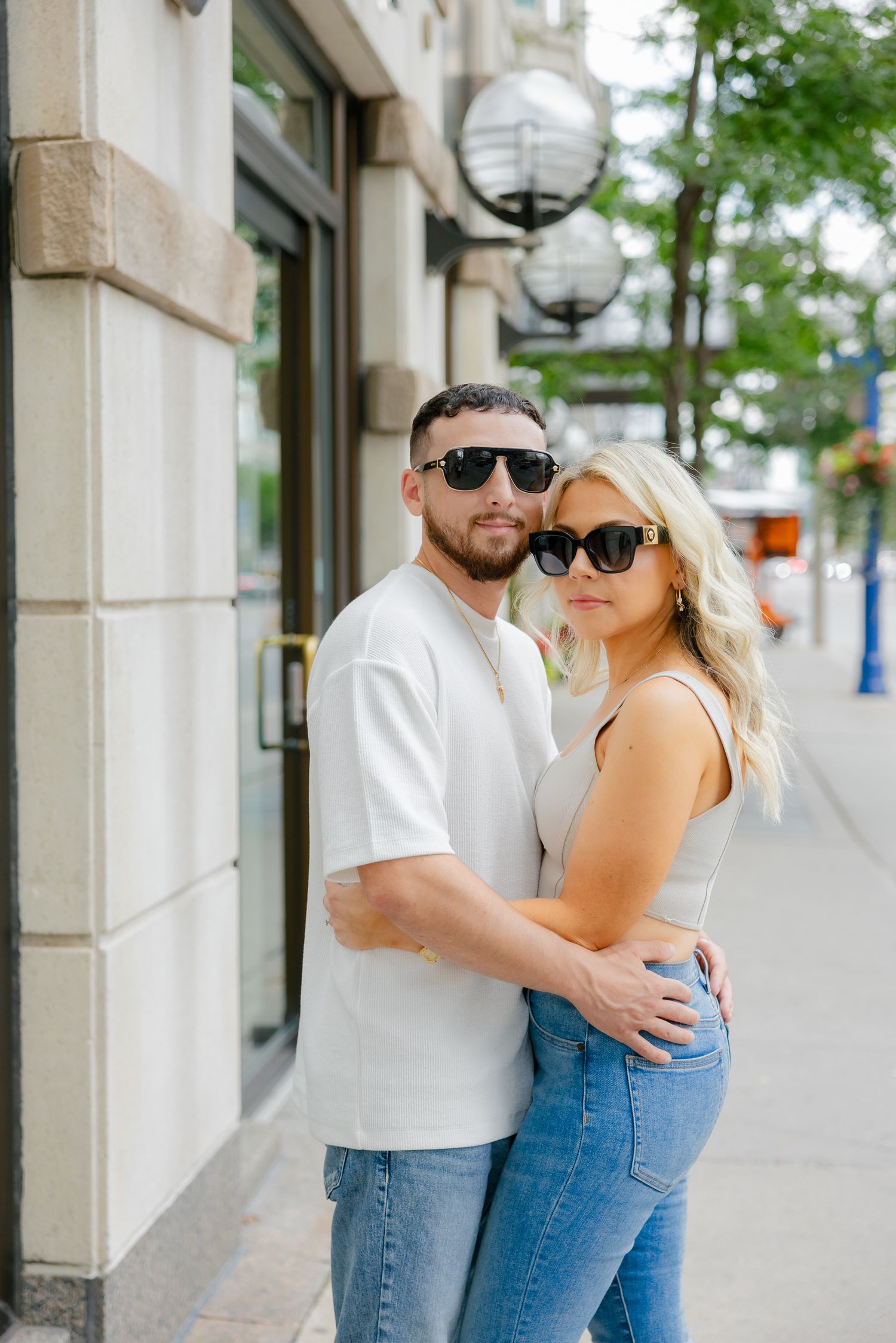 A couple in sunglasses poses intimately together on an urban sidewalk.