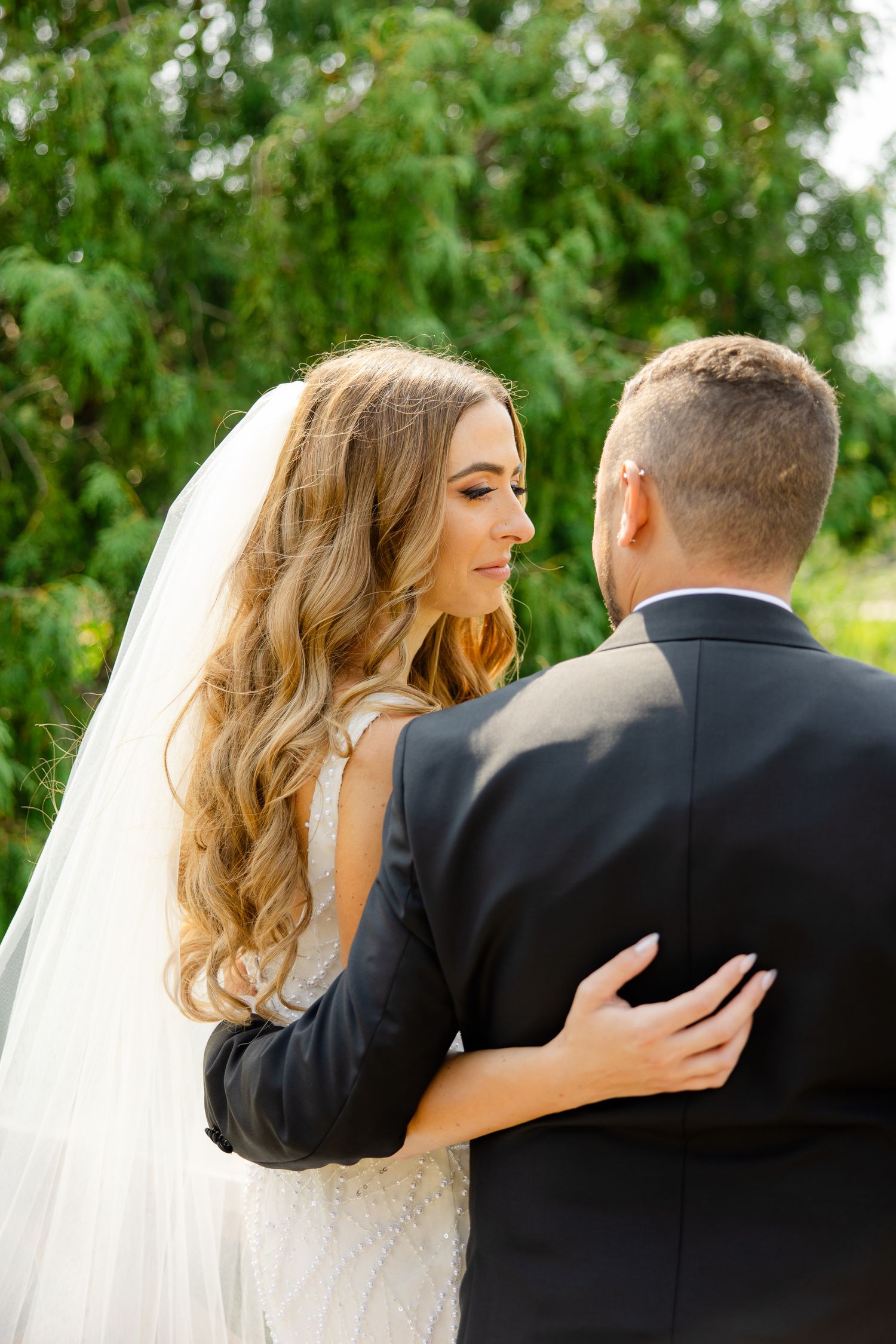 A tender wedding moment captured in a garden as newlyweds embrace, with the bride's flowing veil and lace dress.