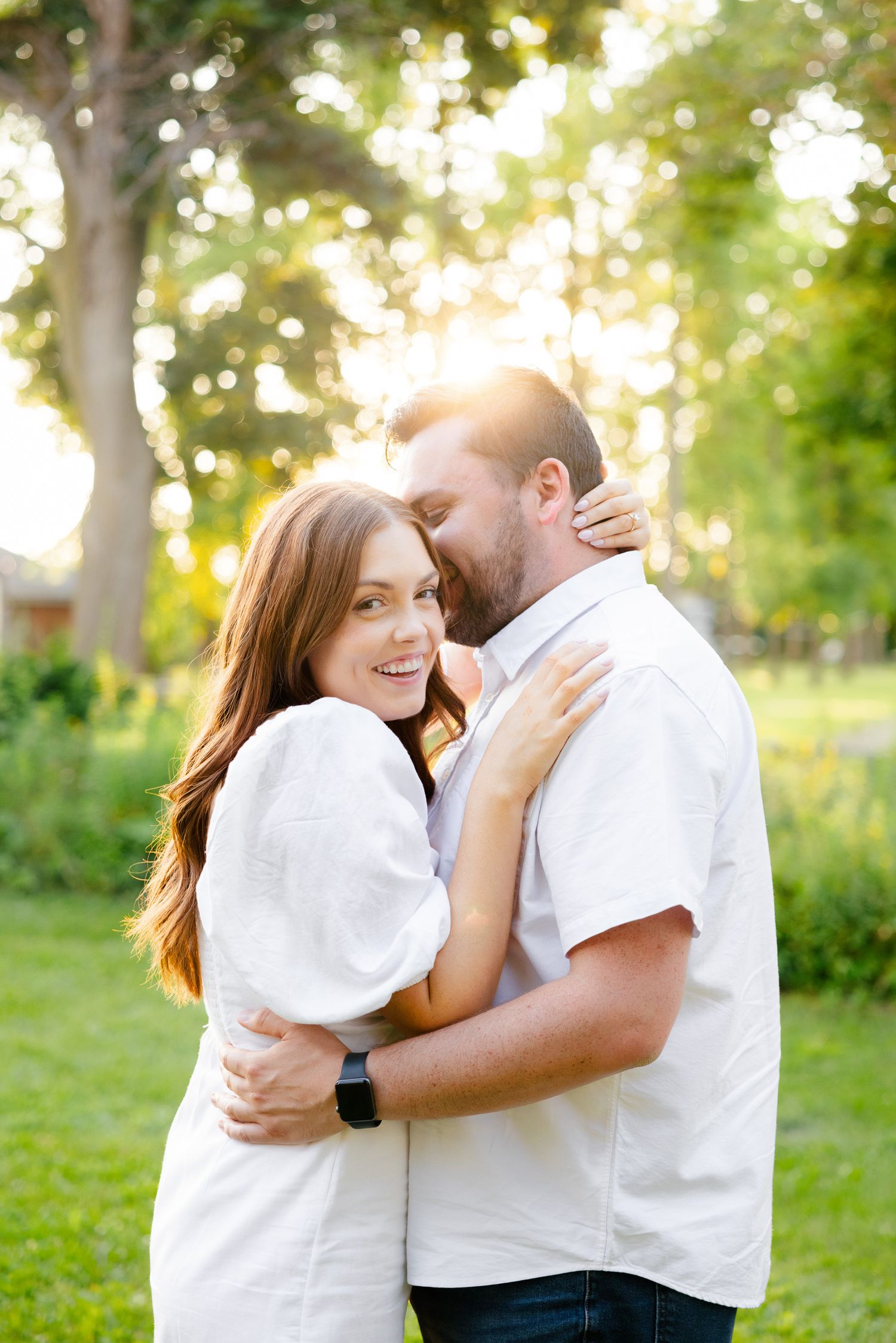 Couple shares intimate moment while embracing outdoors with sunlit trees in background.