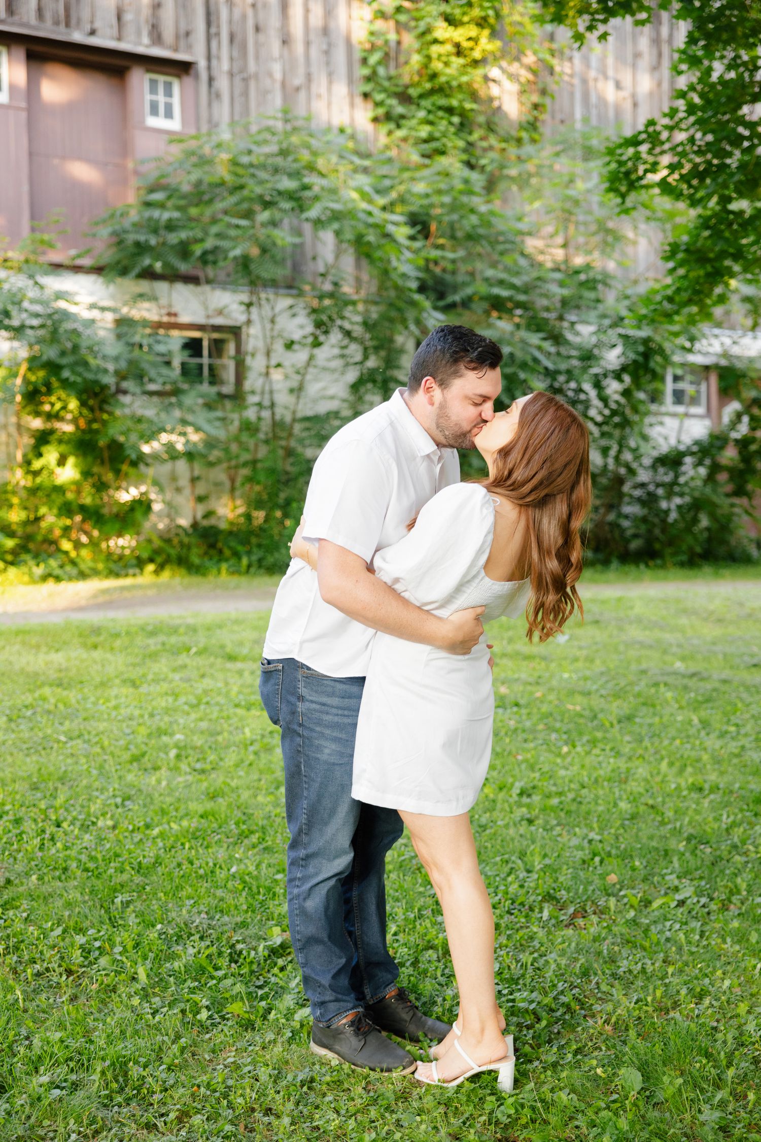 Romantic couple in white sharing an intimate kiss on a green lawn with barn in background.