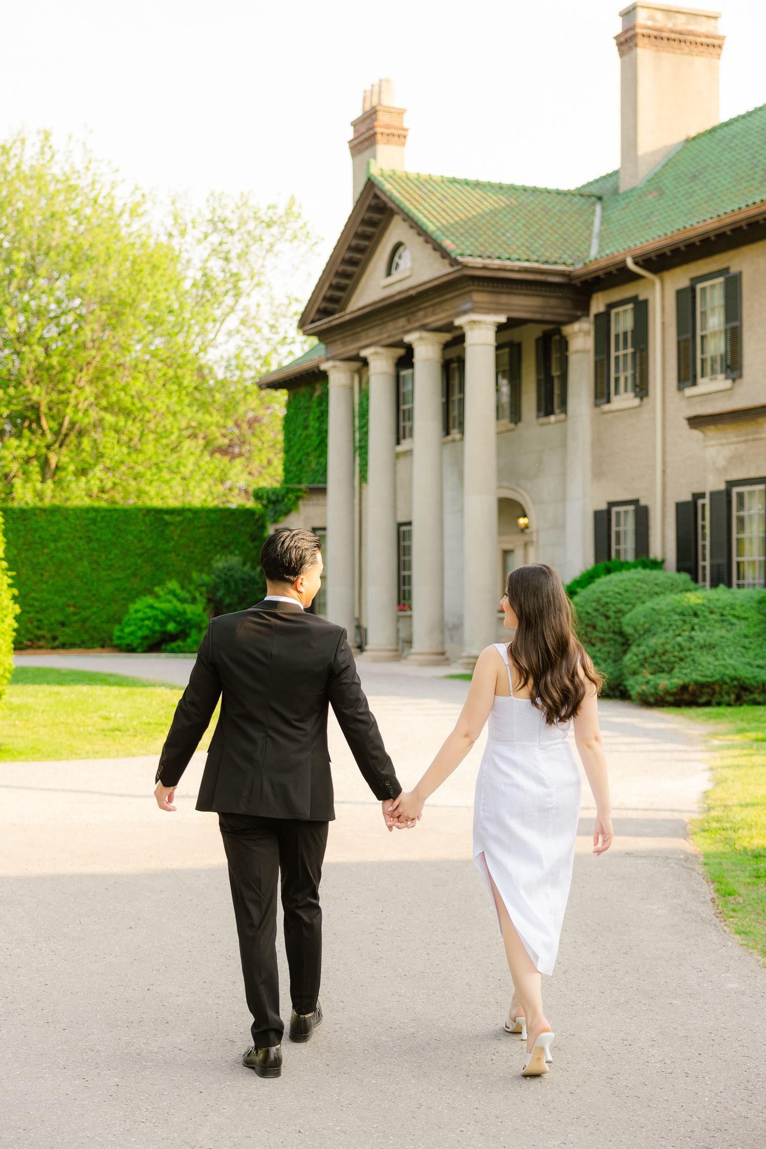 Elegantly dressed couple walking hand in hand toward a grand historic mansion with columns.