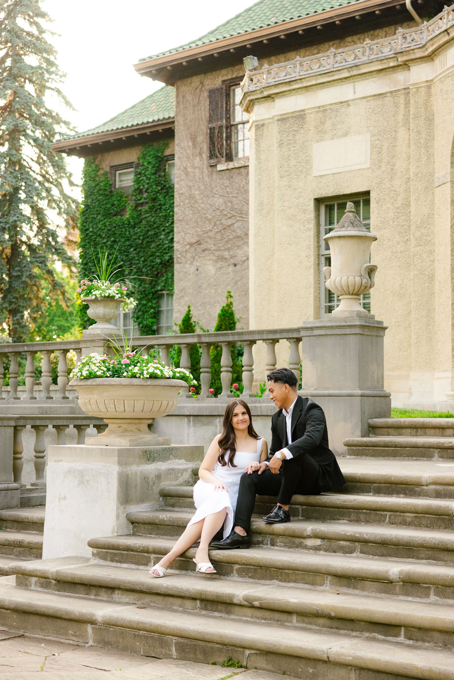 Two people in formal attire sit together on stone steps beside decorative urns and classical architecture.