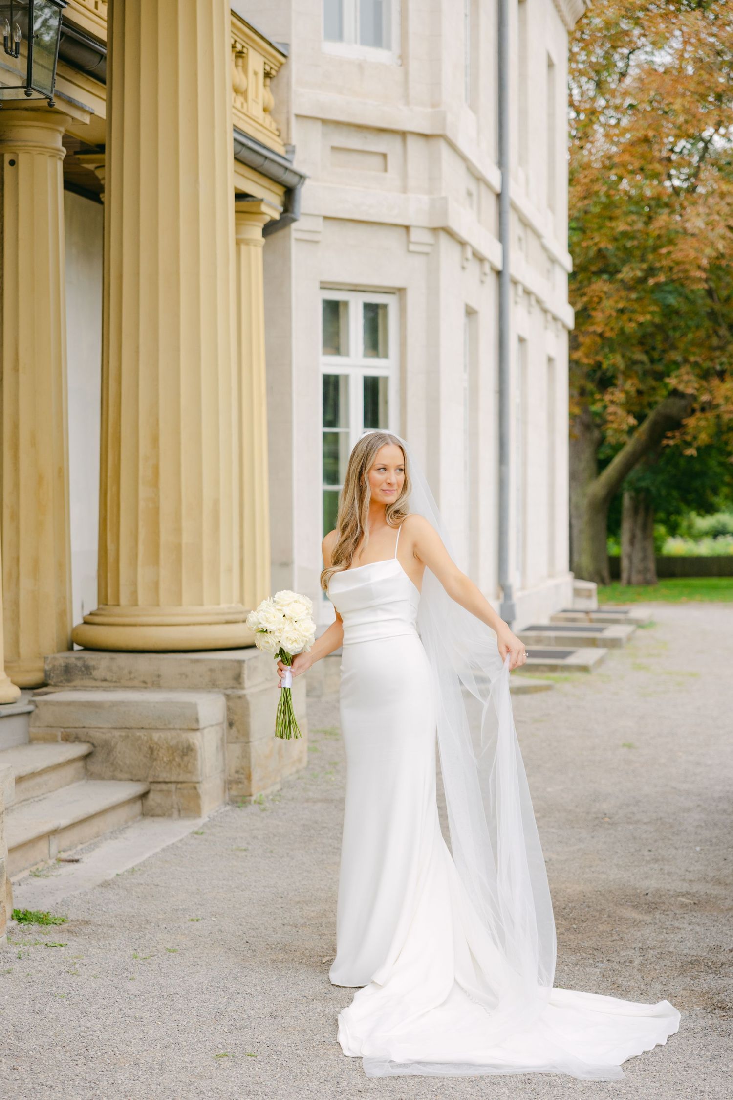 Bride in fitted white wedding dress holds bouquet while posing by ornate stone columns.