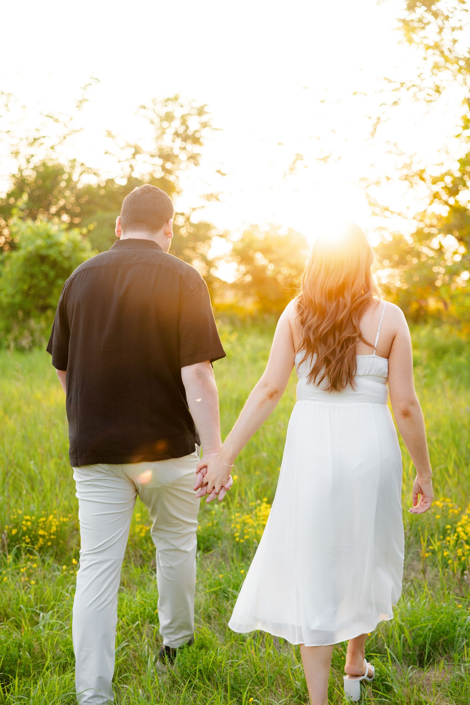 Couple walking hand in hand through tall grass during sunset in countryside setting.