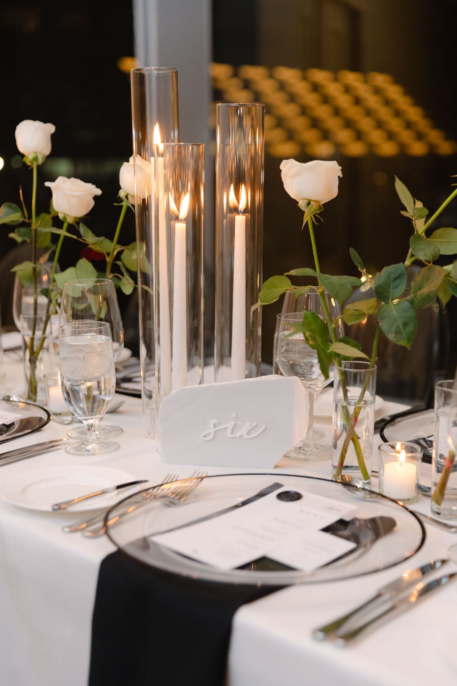 Modern table setting with white roses, tall glass vases, and black and white decor elements.