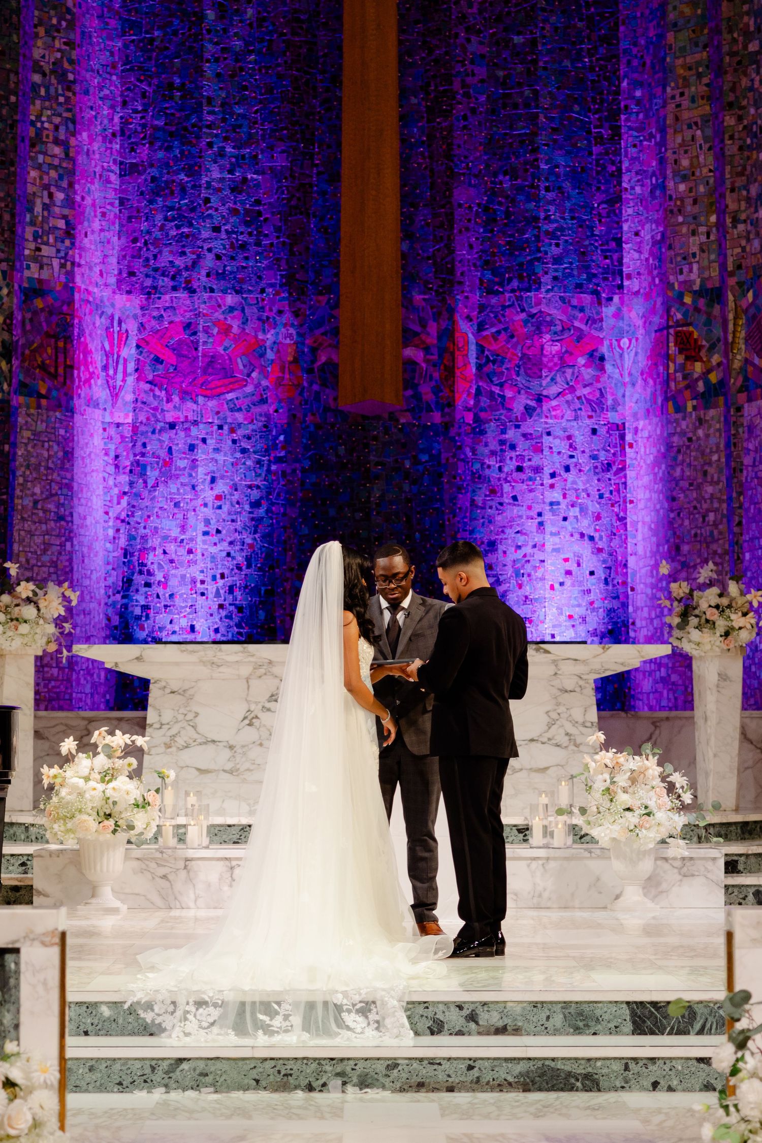 Wedding ceremony at church altar with purple uplighting and white floral arrangements on marble steps.
