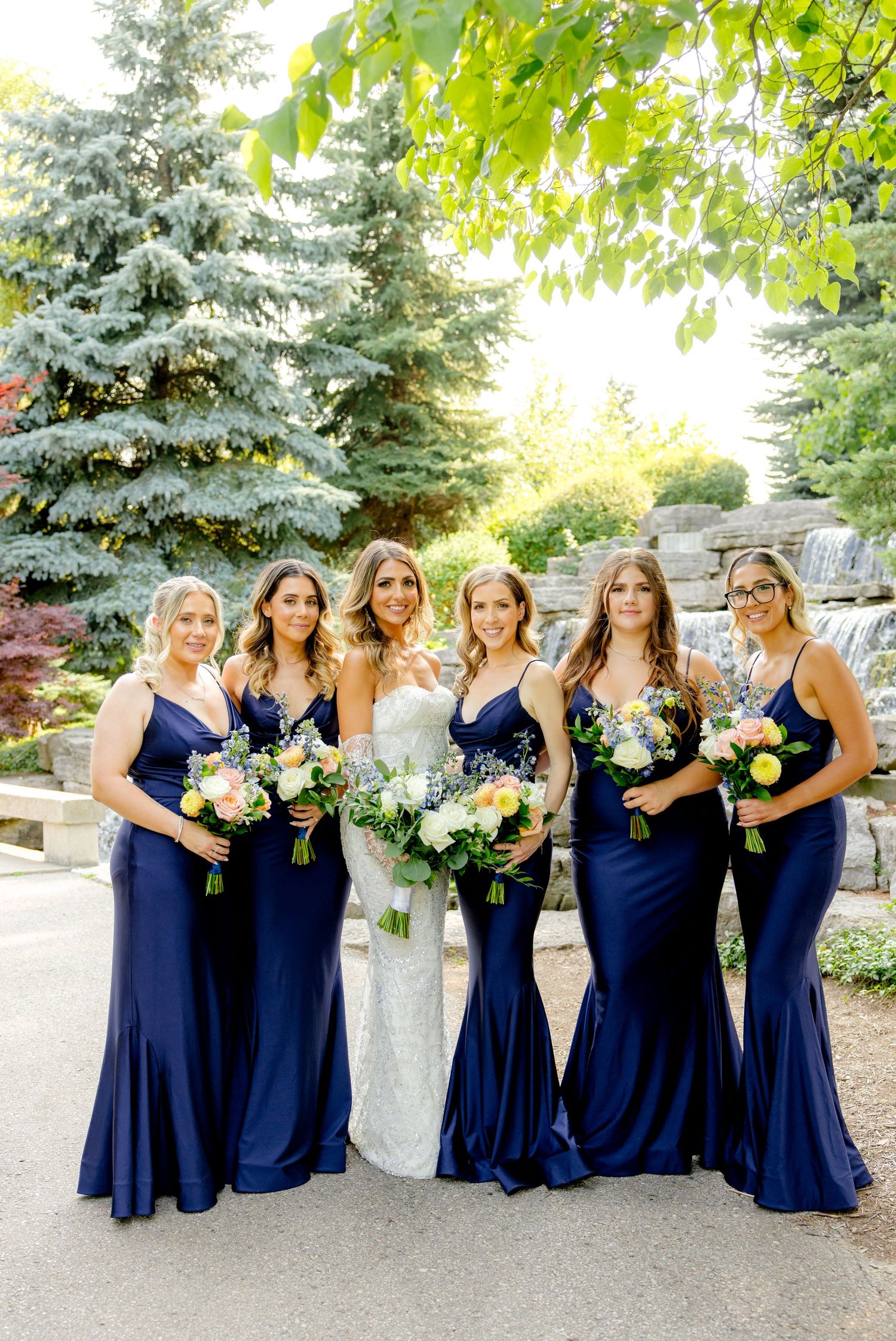 Bridesmaids in navy blue gowns pose with bride holding floral bouquets by waterfall.