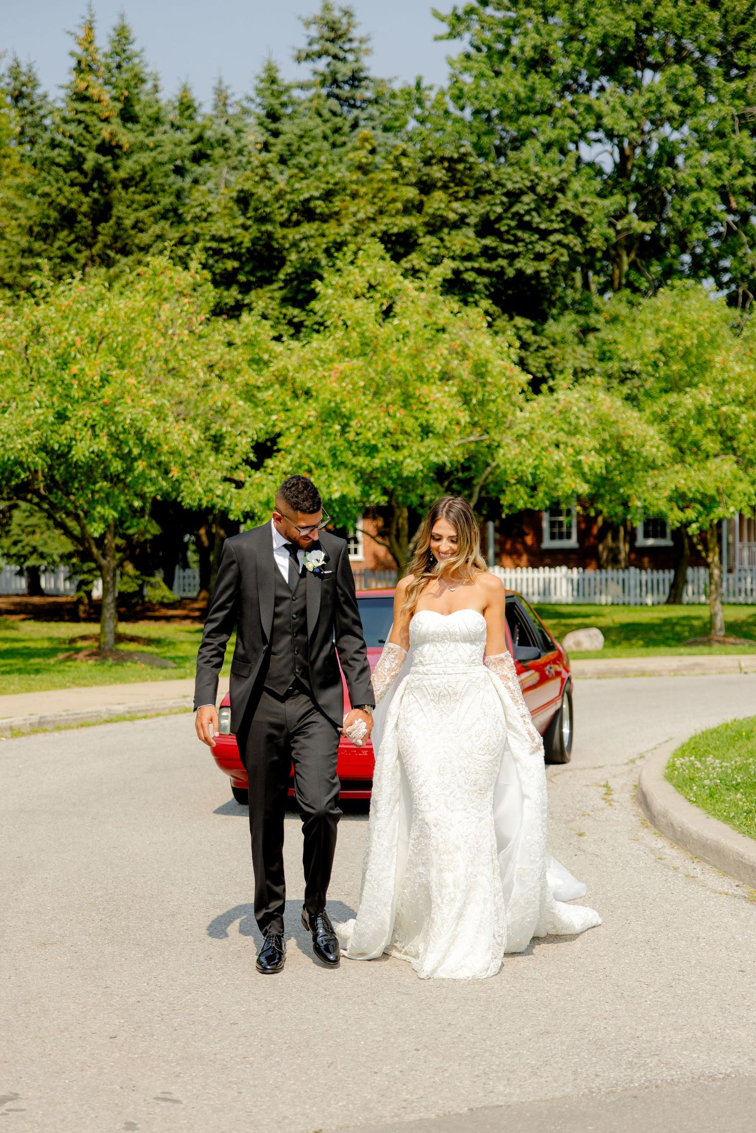 Wedding couple takes a romantic walk together down a tree-lined pathway near their red car.
