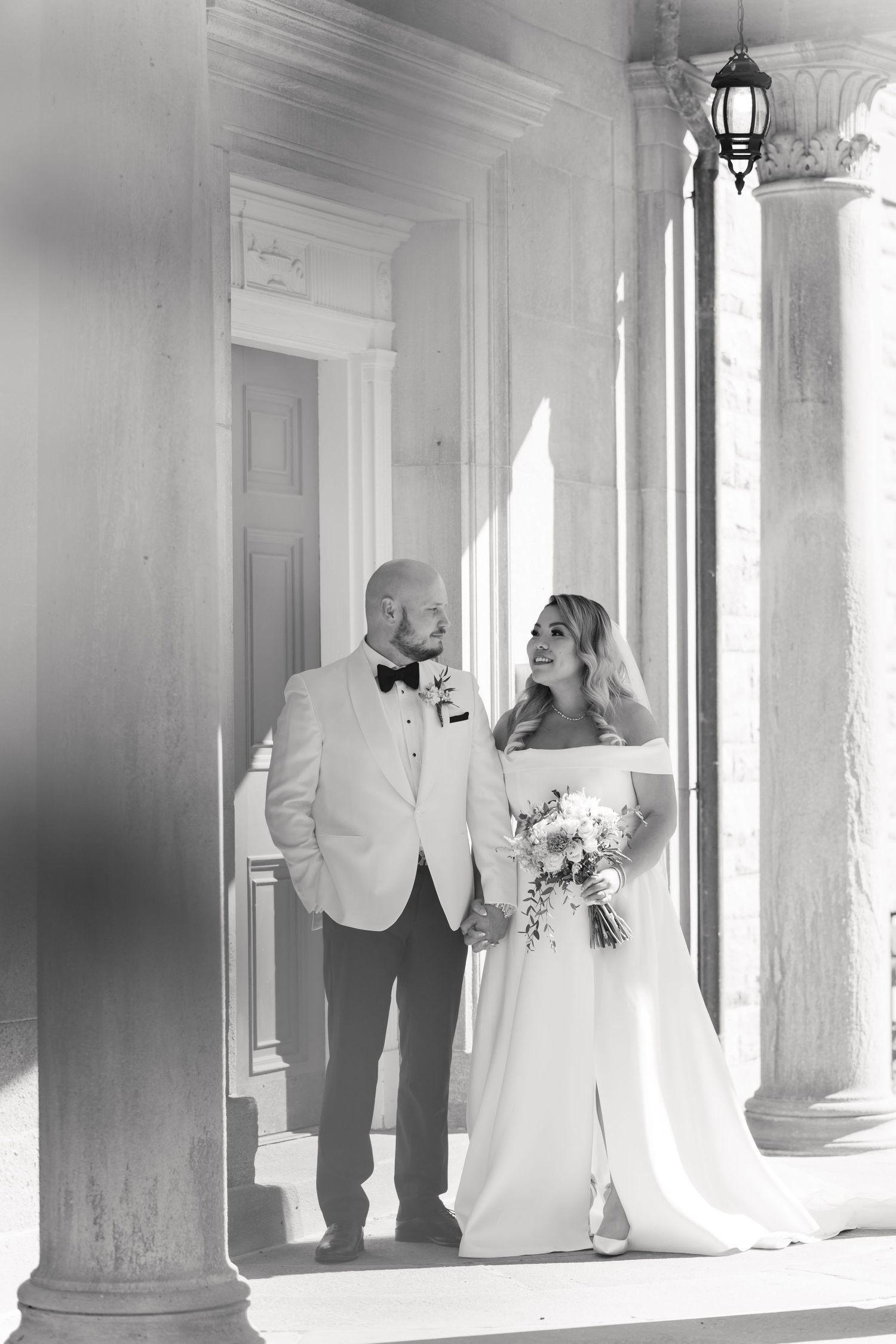 A black and white wedding photo showing a couple standing between classical columns in natural light.