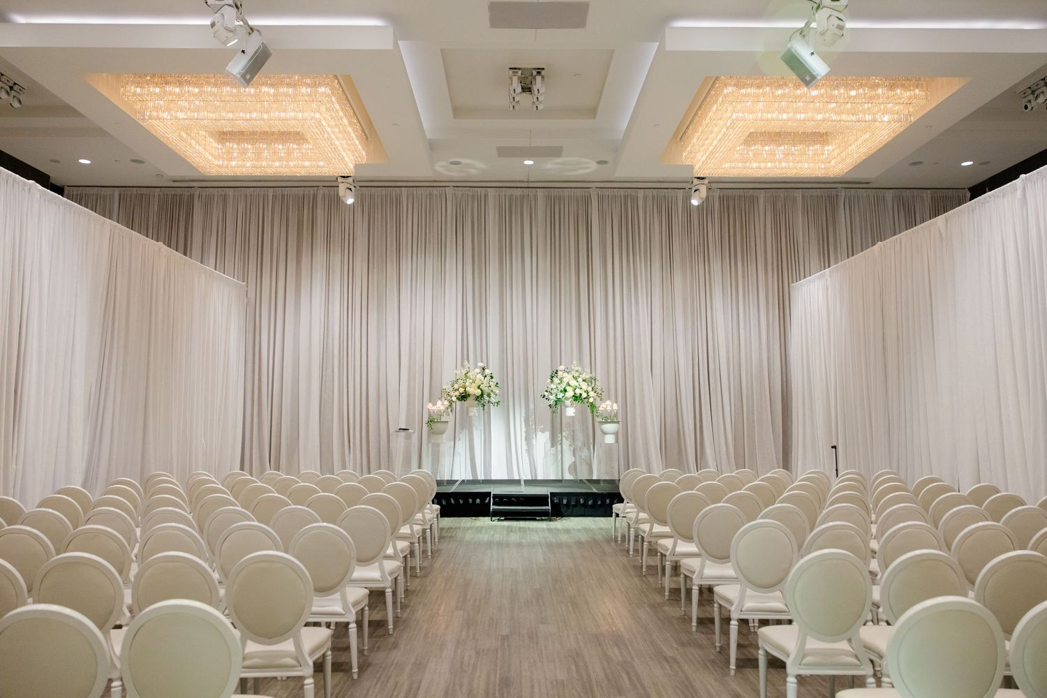 Modern wedding ceremony space with white draping, chandelier lighting and rows of white chairs.