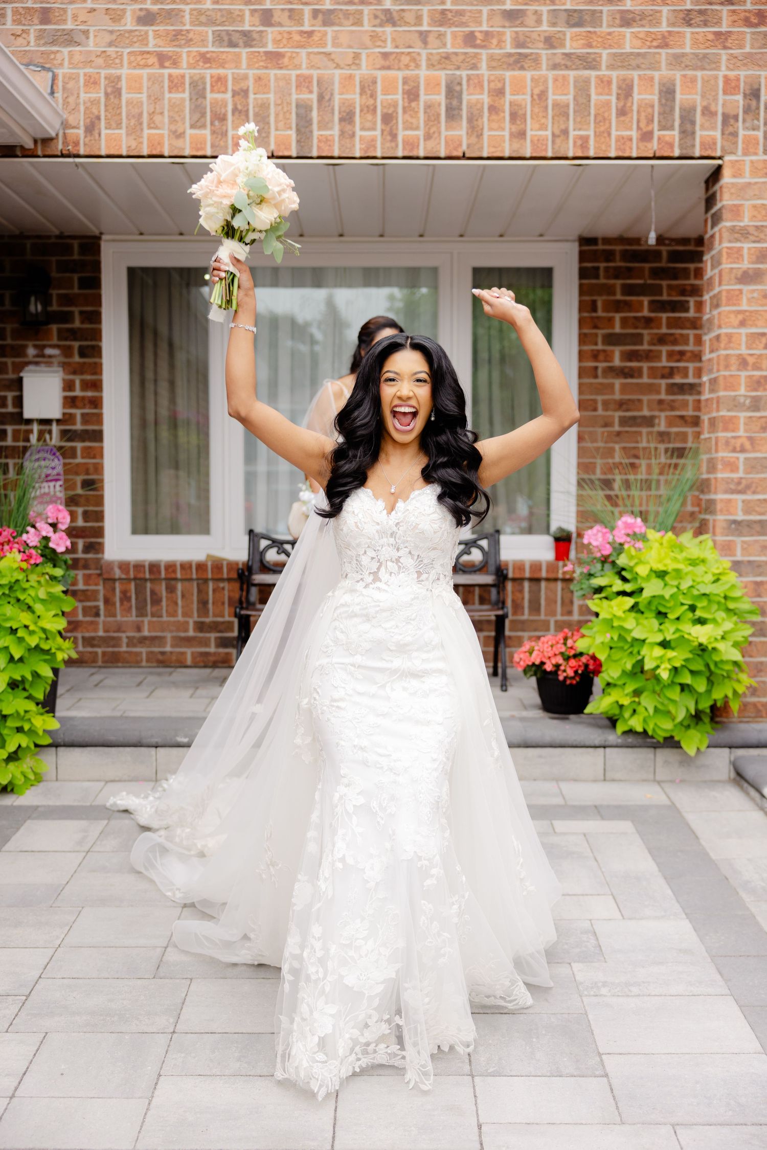 Joyful bride in white lace gown raises bouquet celebrating on stone patio.