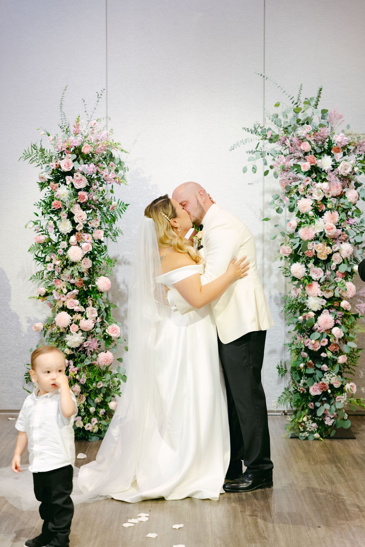 Romantic wedding kiss under floral arch with small child in foreground.