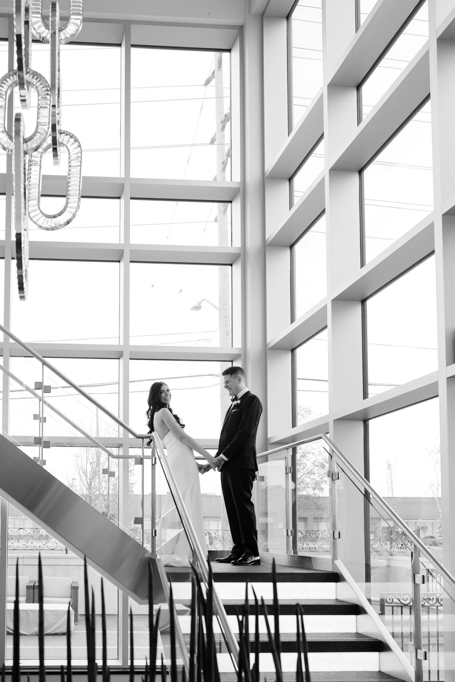 Wedding couple stands together on modern staircase with large windows and dramatic lighting.