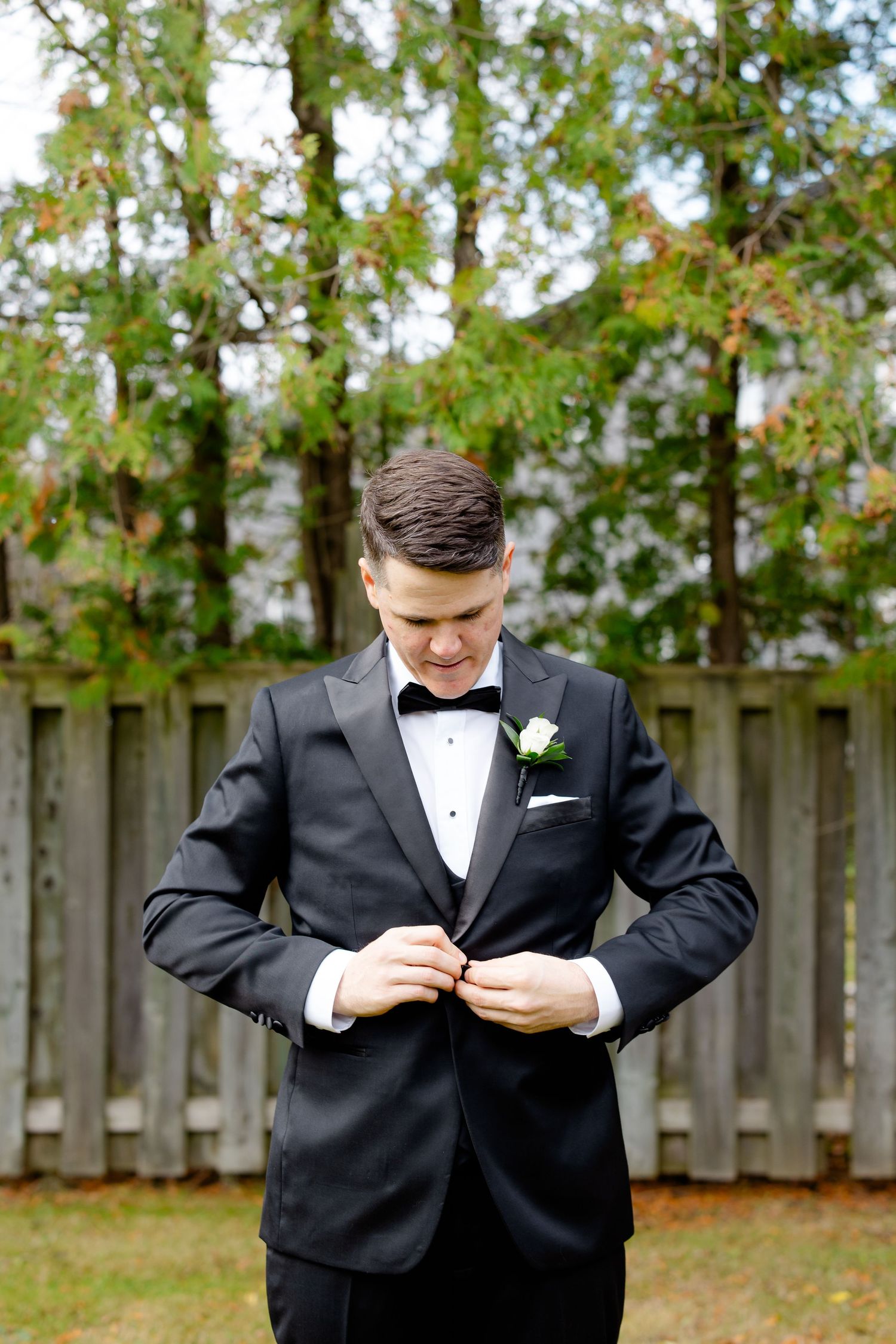 A person in a black tuxedo adjusts their jacket while standing against a wooden fence background.
