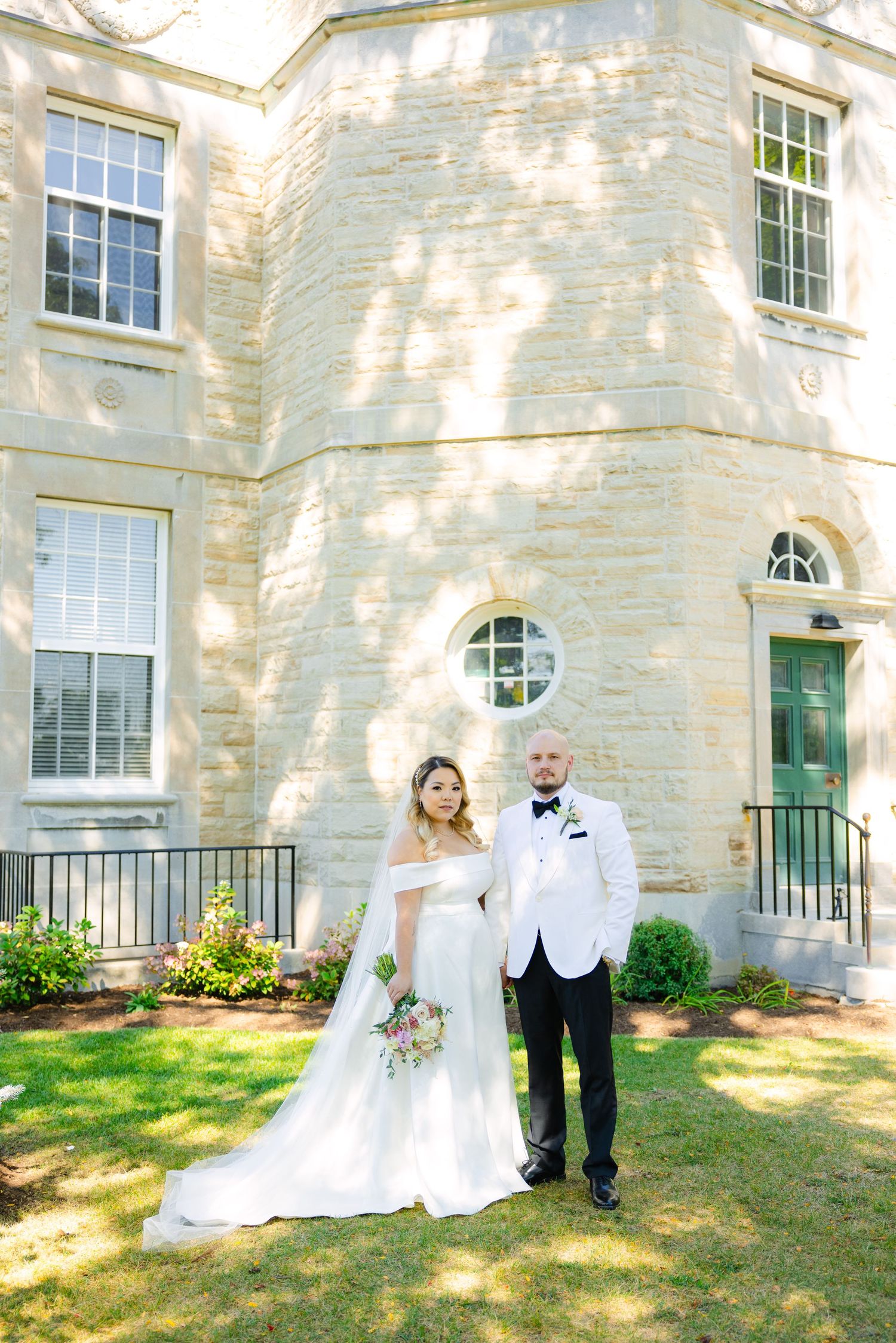 The wedding couple stands in elegant attire before a grand stone building with round windows.