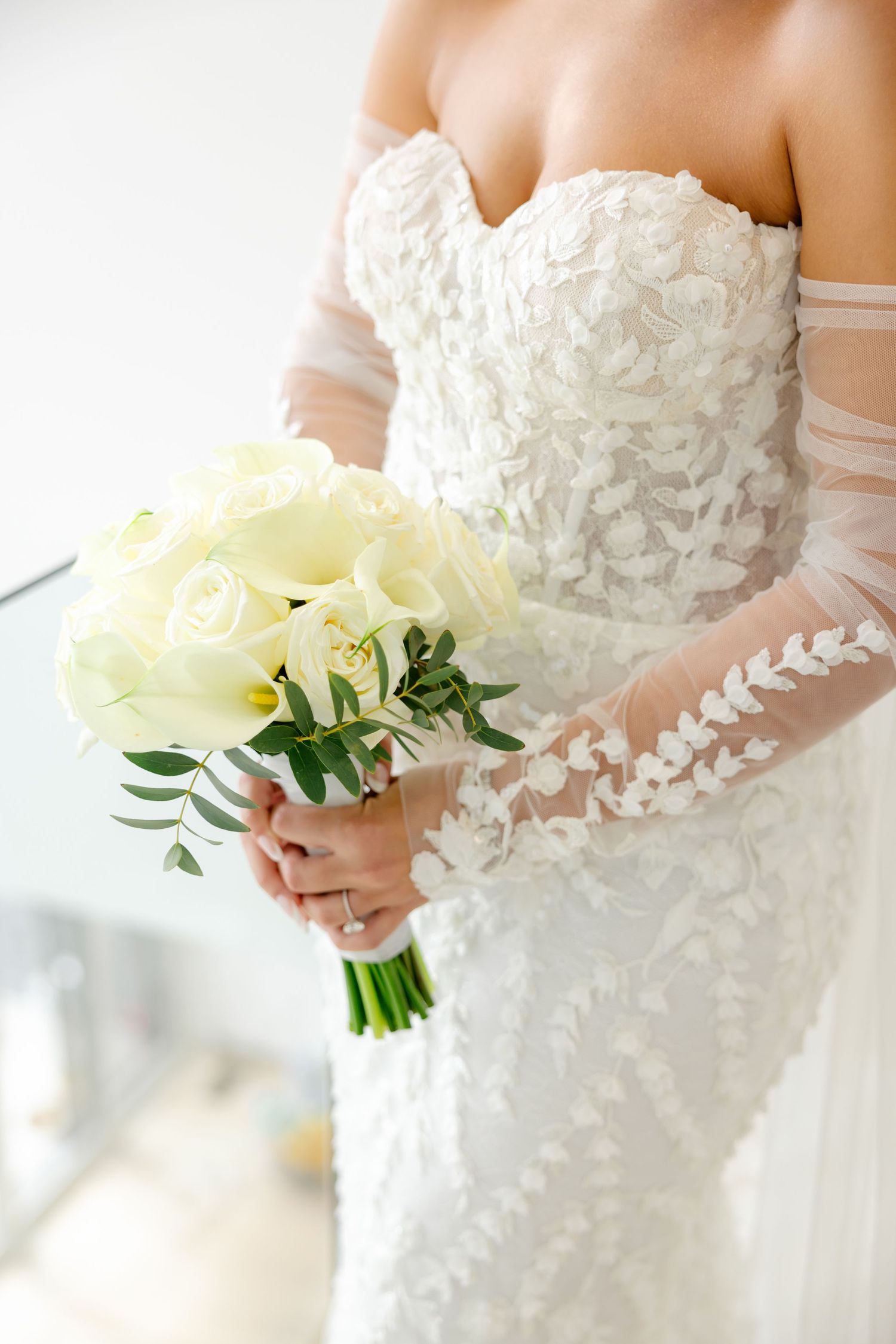 Close-up detail of lace wedding dress bodice and cream rose bouquet with greenery.