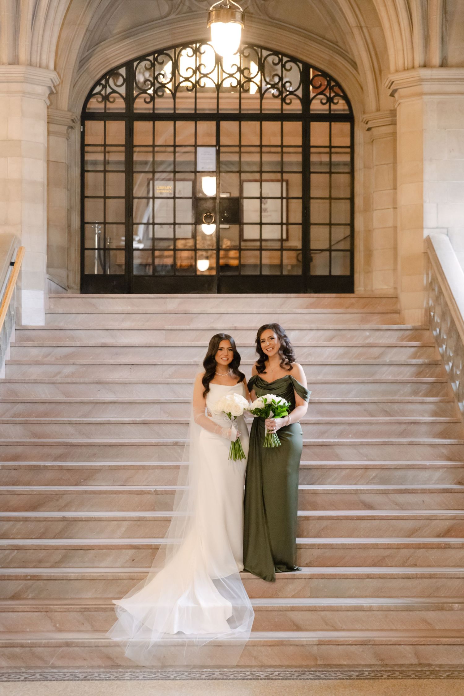 Two figures in wedding attire pose on marble staircase with ornate gothic window and pendant lighting above.