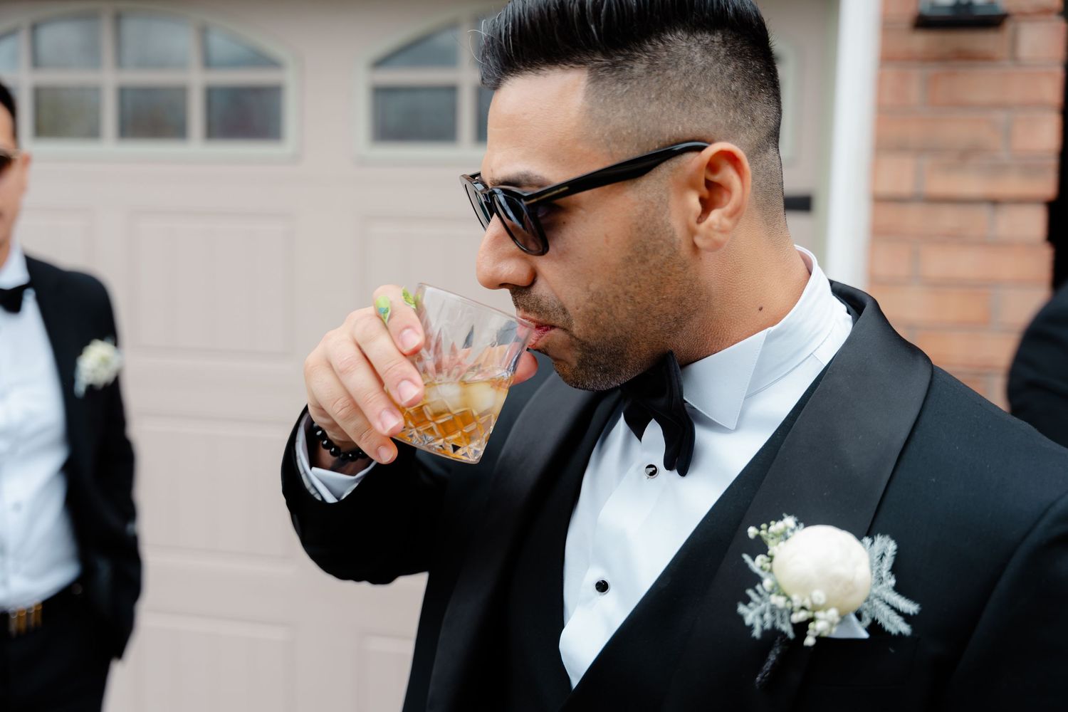 Groomsman in formal black tuxedo and sunglasses drinking from a glass outdoors.