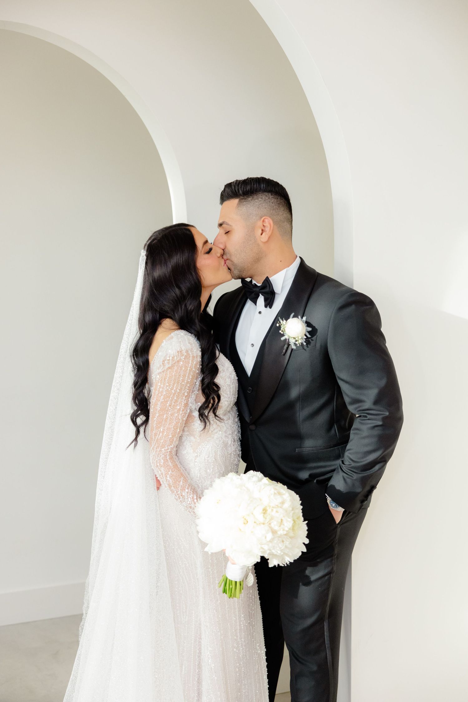 Romantic wedding couple sharing intimate moment under white archway with bridal bouquet.