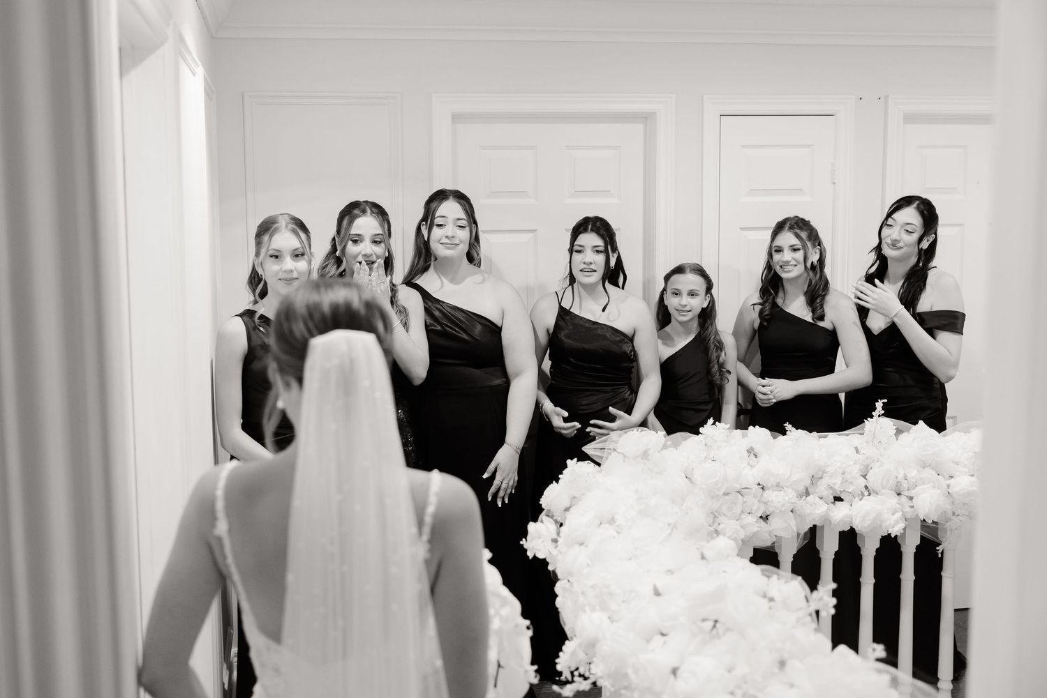 A black and white wedding photo captures a candid moment with bridesmaids in elegant black dresses gathered in a room.