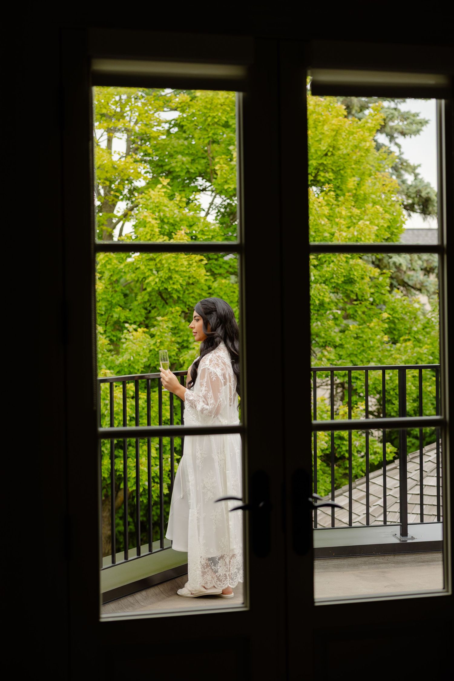 View through window frame of person in white dress standing on balcony surrounded by lush green trees.
