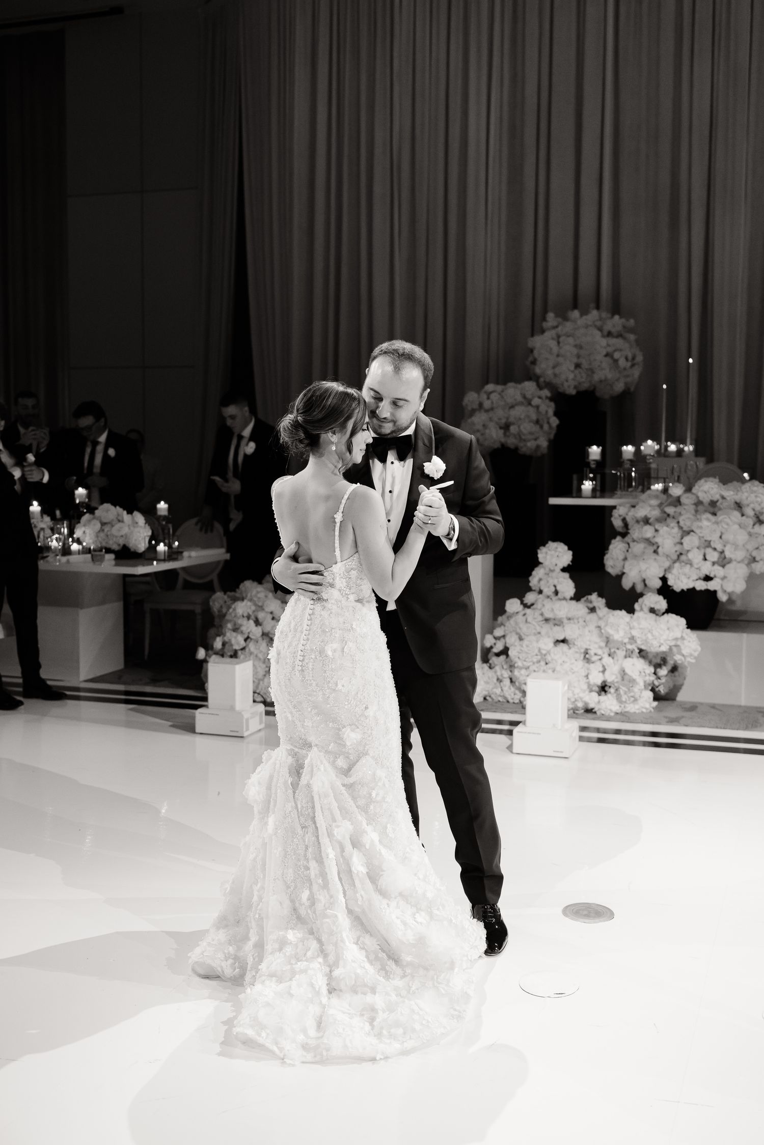 A couple shares their first dance at an elegant wedding reception with white floral decor and candlelight.