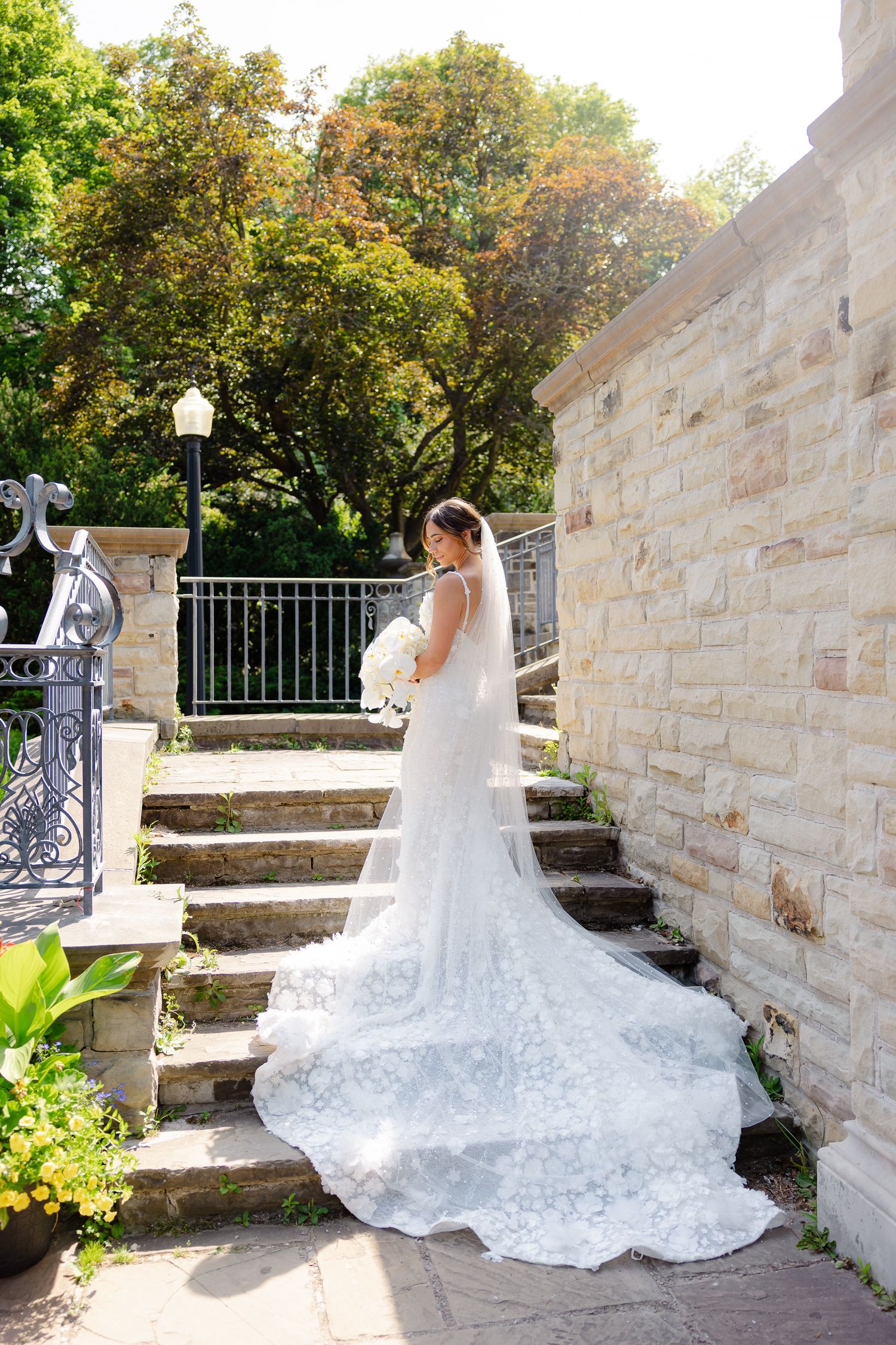 Long lace train wedding dress showcased in natural sunlight by brick wall and stone steps with wrought iron railing.