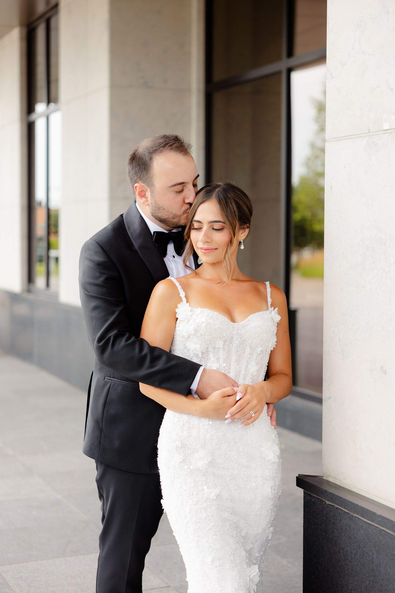 A bride in a white lace gown and groom in black tuxedo share an intimate moment together outside a building.