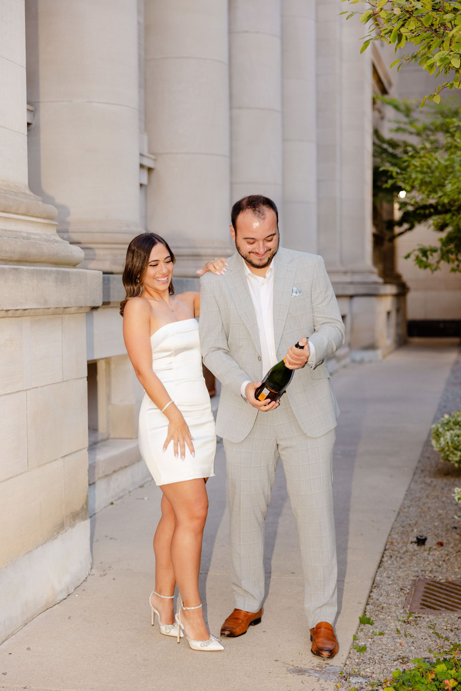 Couple in formal wear stands by architectural columns with champagne.