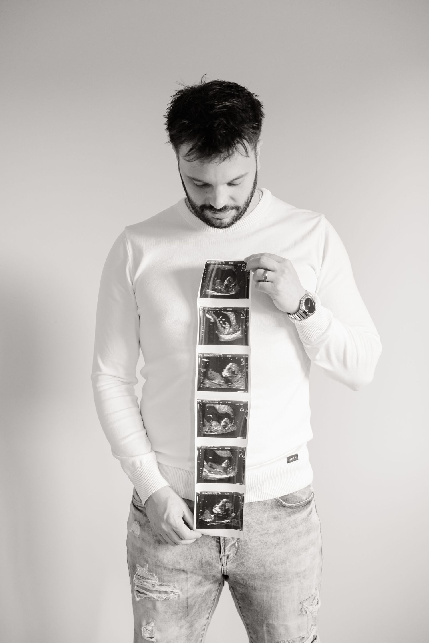 Man in white sweater holds strip of ultrasound images, looking down at them with a smile.